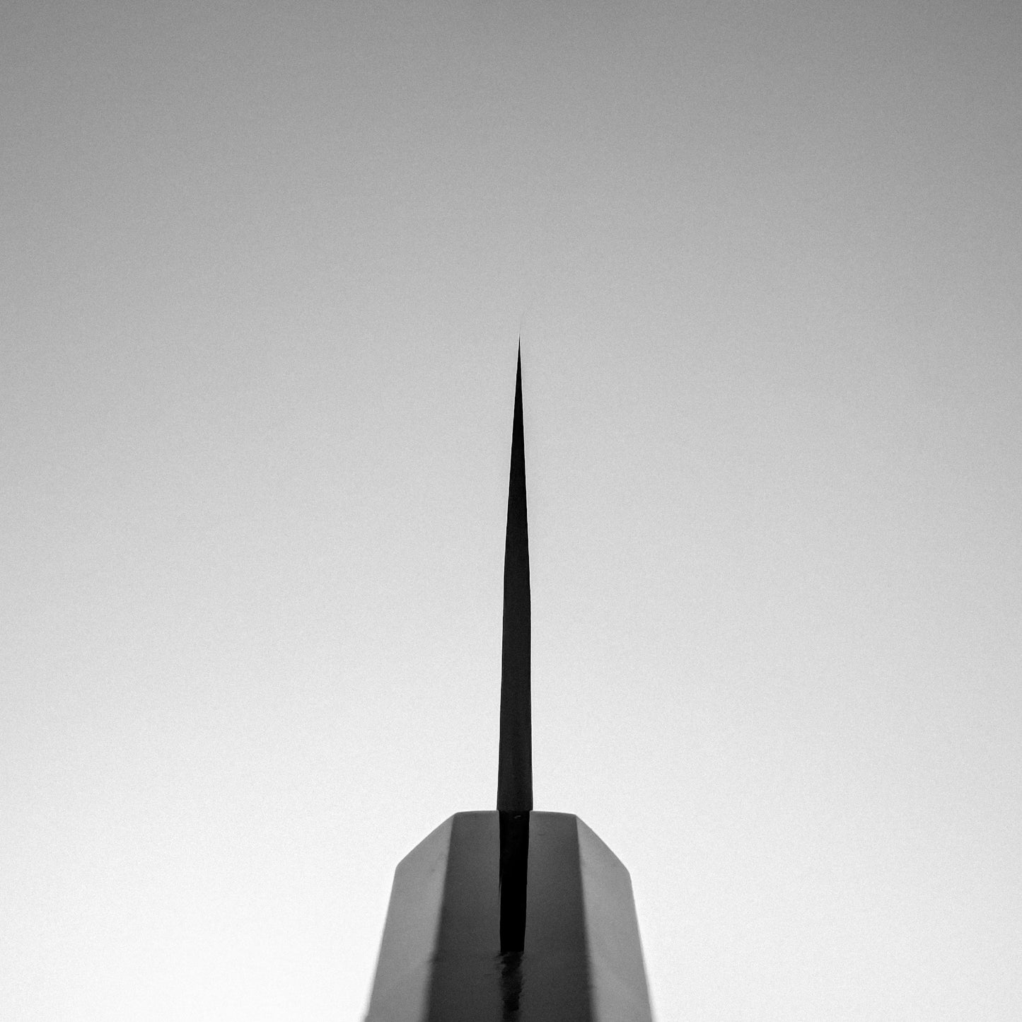 A sharp, narrow spire rises from a geometric base against a pale gray sky, evoking the precise symmetry and minimalist allure of the Yoshikane Hatsukokoro x Yoshikane nashiji SKD nakiri 165mm knife.