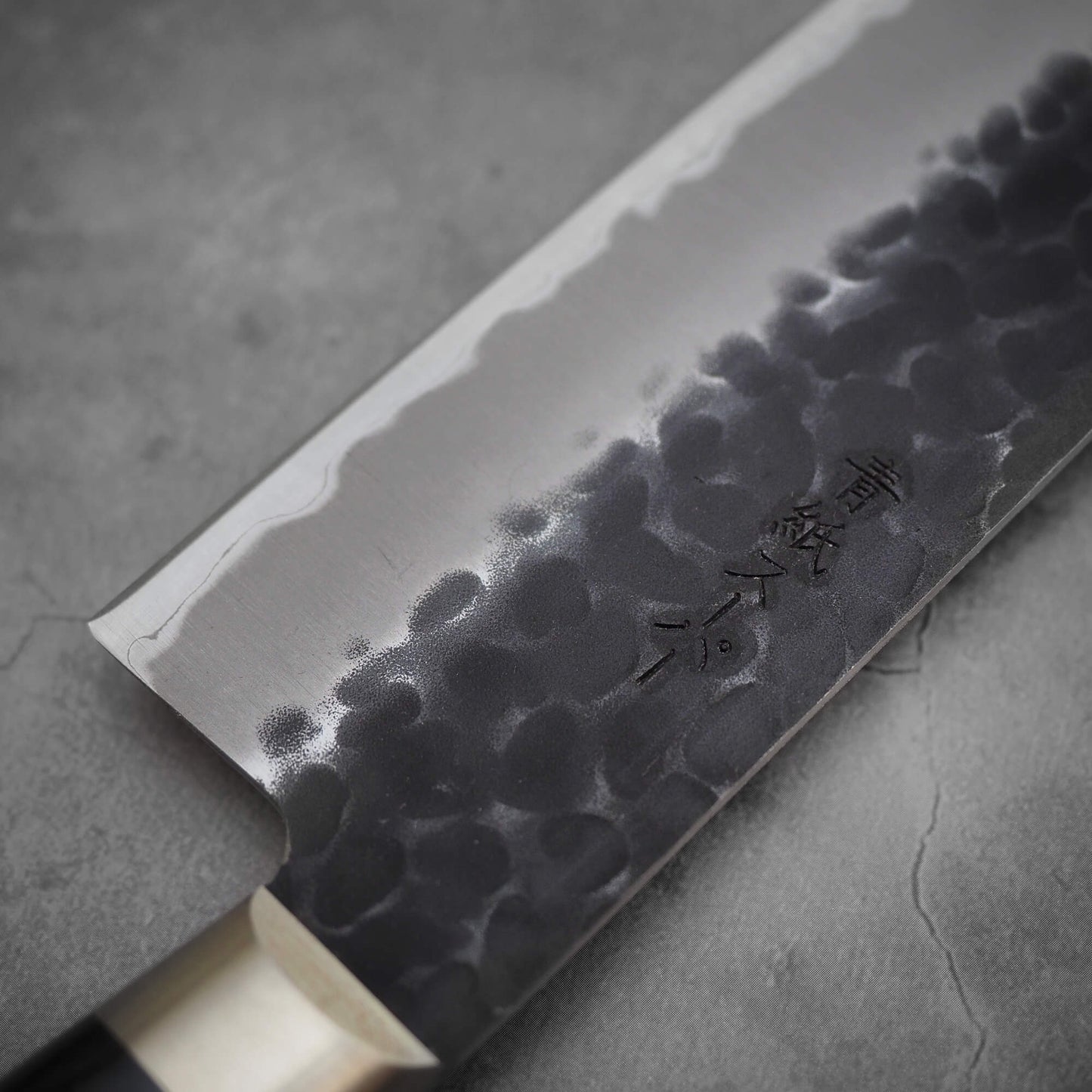 Close up view of Ittosai Kotetsu tsuchime kurouchi aogami super kiritsuke gyuto. Image focuses on the left side of the blade showing the kanji