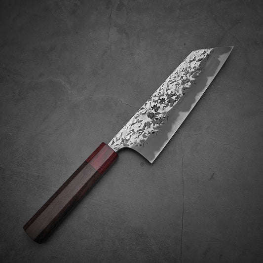 The Kisuke Manaka tsuchime white#2 bunka 170mm knife by Kisuke Manaka, featuring a hammered white#2 steel blade and an octagonal rosewood handle, rests on a textured gray surface.