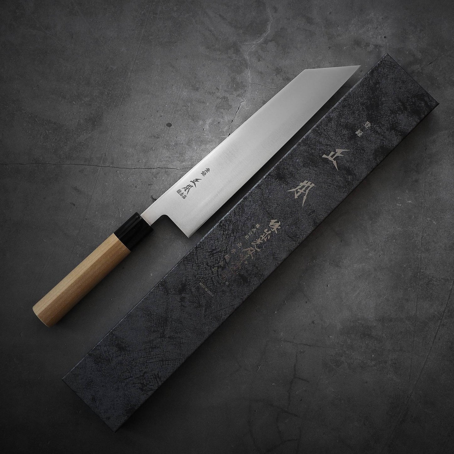 A Masamoto MV kiritsuke gyuto 240mm with a wooden handle lies next to a rectangular black box with Japanese writing, set on a dark gray textured surface.