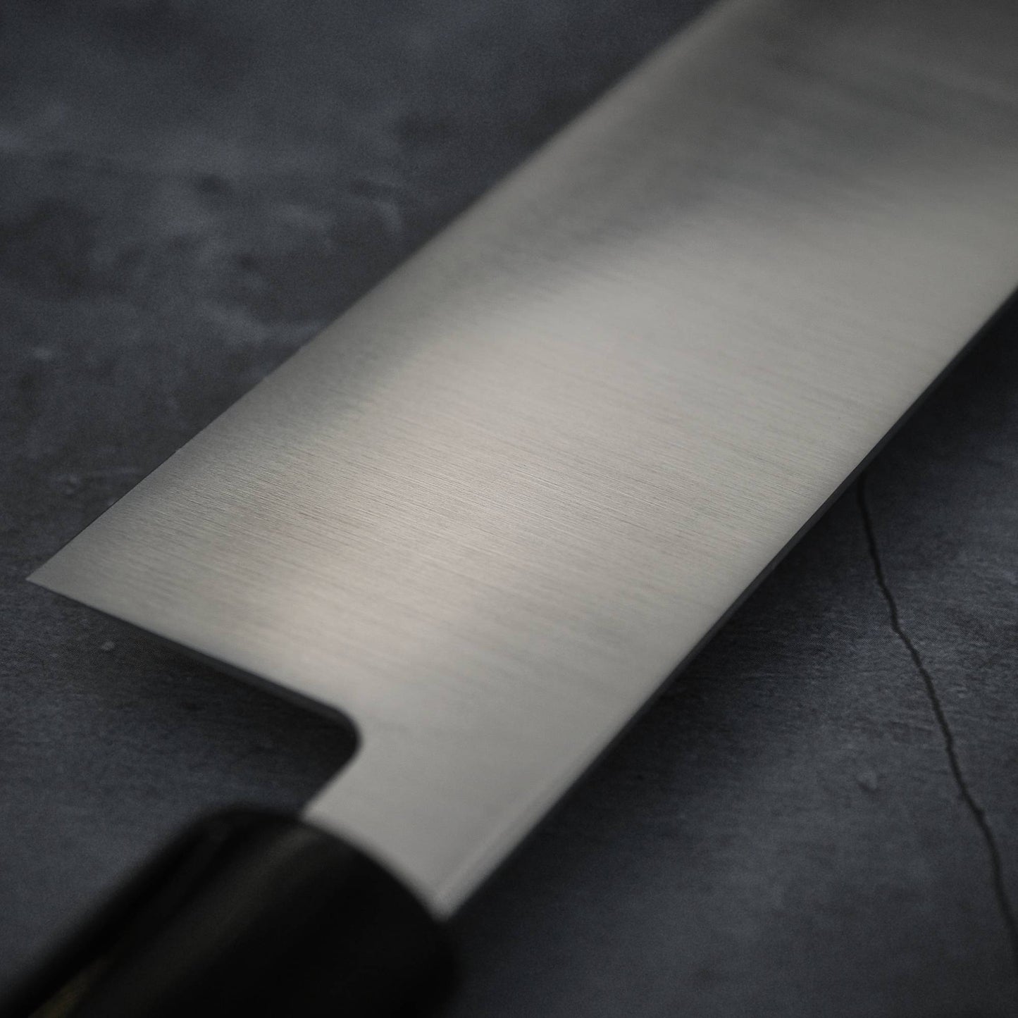 A close-up of the Masamoto MV kiritsuke gyuto 240mm kitchen knife with a black handle, set on a dark textured surface. The blade from Masamoto is stainless steel, clean, and polished.
