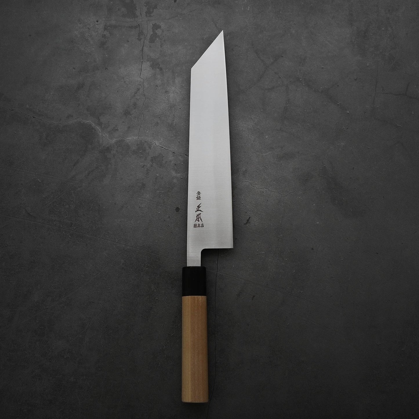 The Masamoto MV kiritsuke gyuto 240mm features an angular MV stainless steel blade with Japanese characters near the wooden handle and rests on a dark, textured surface.
