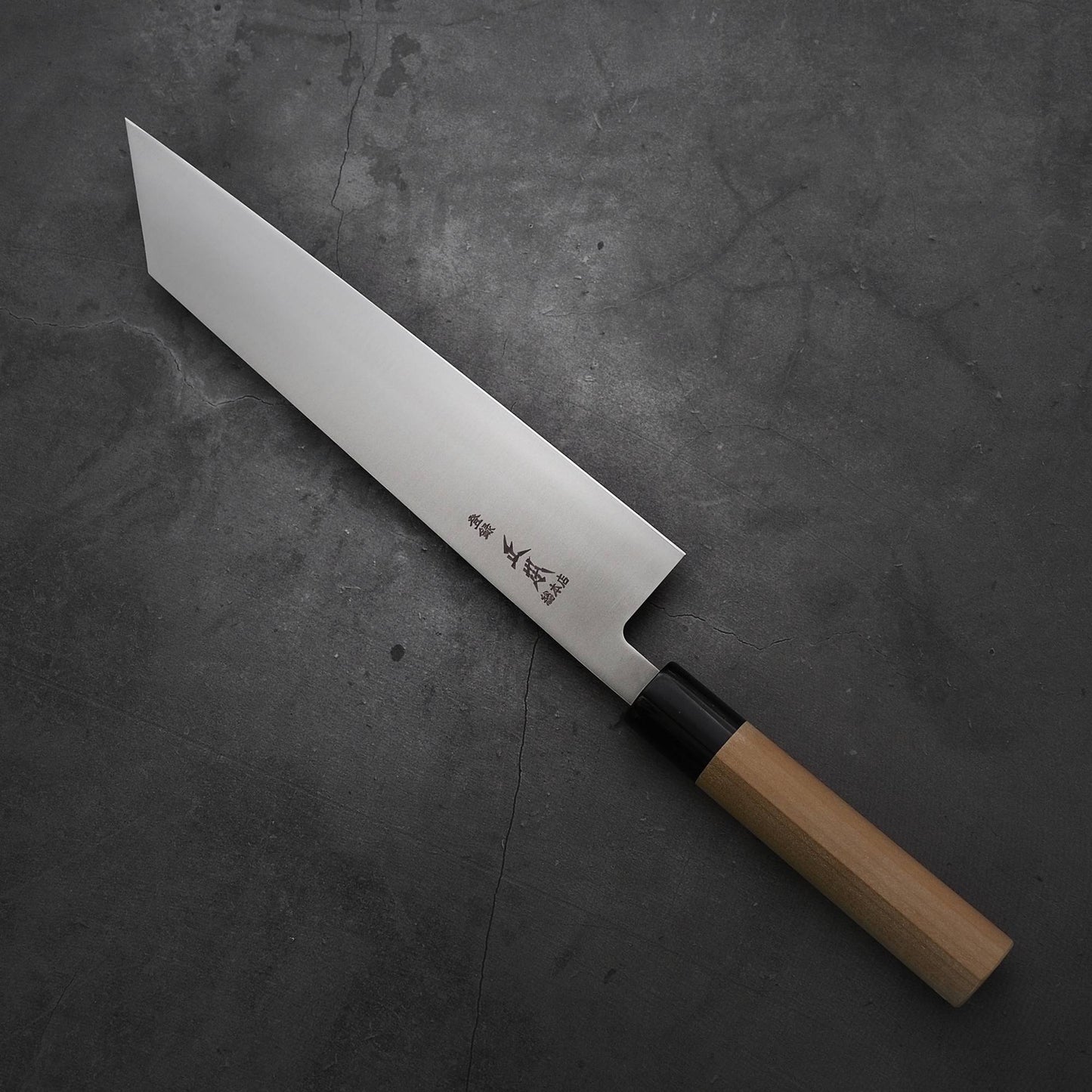 A Masamoto MV kiritsuke gyuto 240mm kitchen knife with a wooden handle and long, angular MV stainless steel blade rests on a dark, textured surface. Japanese characters are engraved near the base of the Masamoto blade.