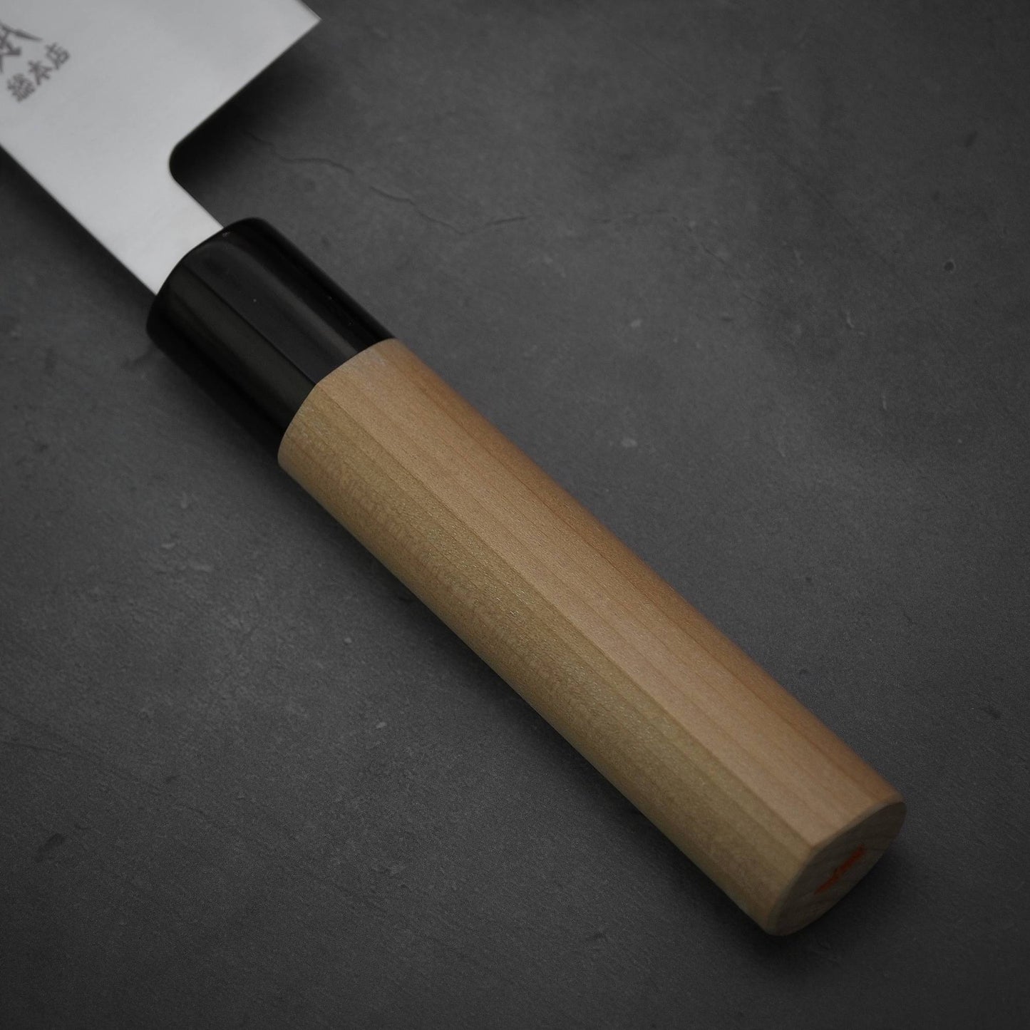 Close-up of the Masamoto MV kiritsuke gyuto 240mm, featuring a wooden handle and black bolster, by Masamoto. The MV stainless steel blade is partially visible as it rests on a dark gray textured surface.