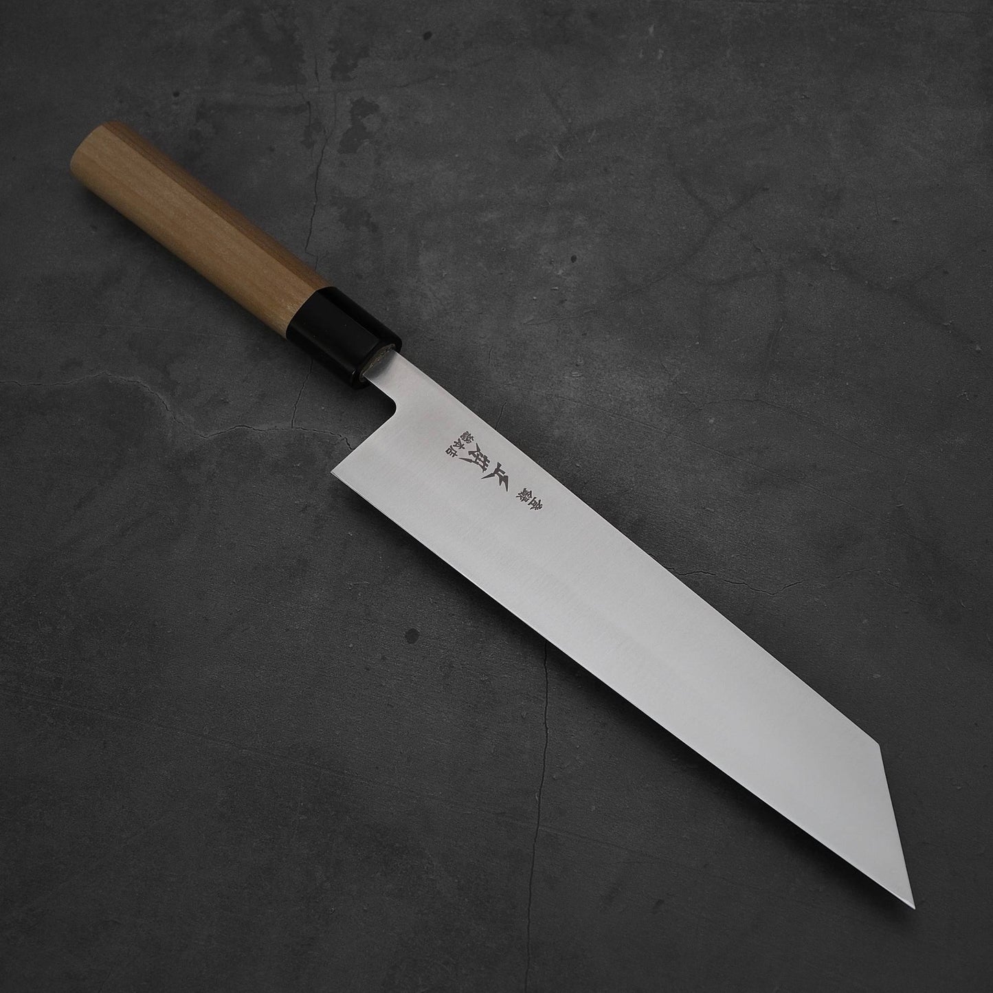 The Masamoto MV kiritsuke gyuto 240mm, a Japanese-style knife with a wooden handle and straight-edged MV stainless steel blade engraved with kanji, rests on a dark, textured surface.