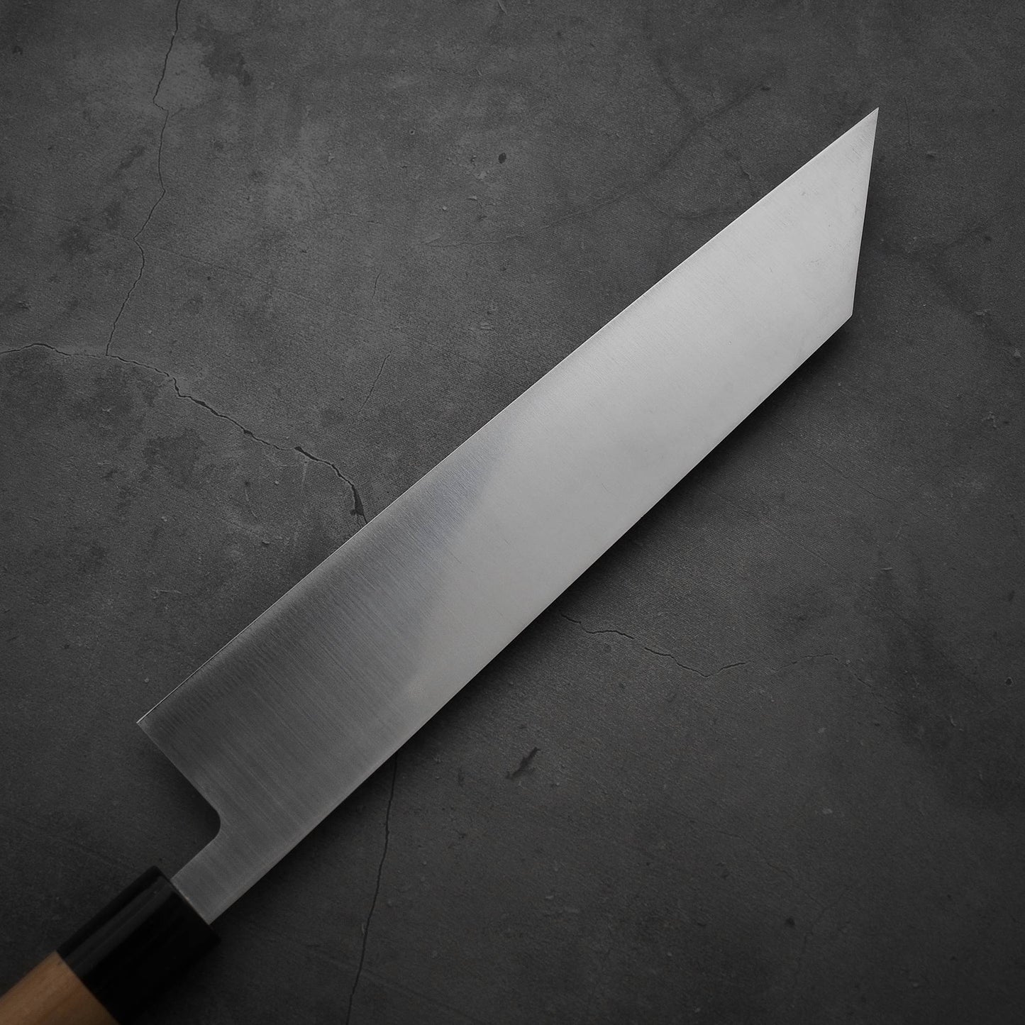 The Masamoto MV kiritsuke gyuto 240mm, a Japanese chefs knife by Masamoto, has a rectangular wooden handle and a long MV stainless steel blade with an angled tip, displayed on a dark, textured cracked surface.