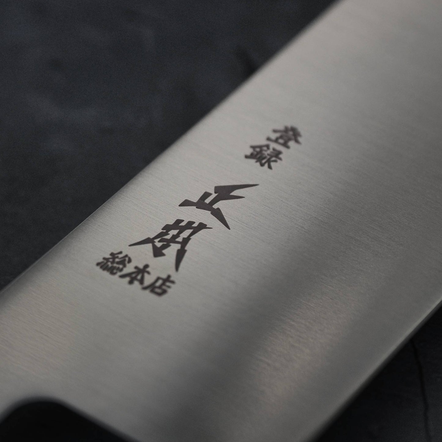 Close-up of the Masamoto MV kiritsuke gyuto 240mm blade in MV stainless steel, engraved with Japanese characters, by Masamoto, photographed against a dark background.