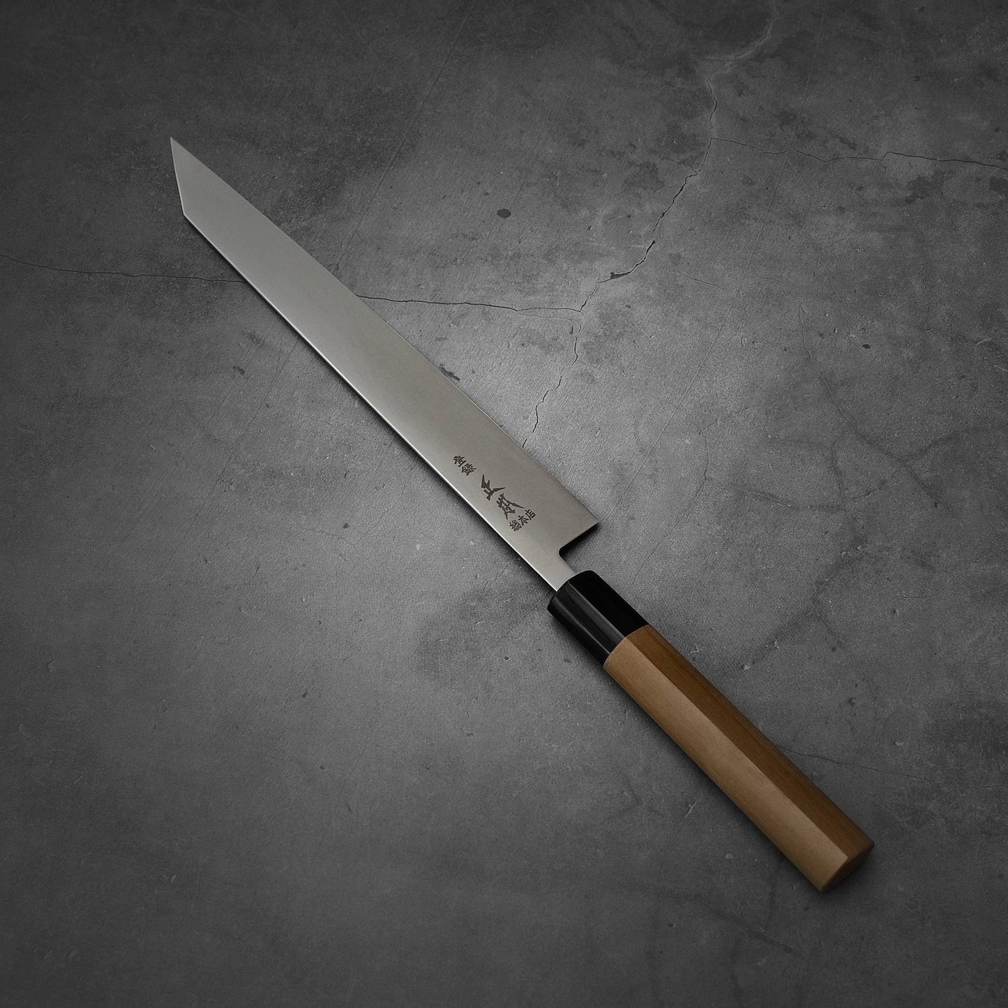 A Masamoto MV kiritsuke sujihiki 255mm knife with a long, pointed MV stainless steel blade and wooden handle is displayed on a dark, textured surface.