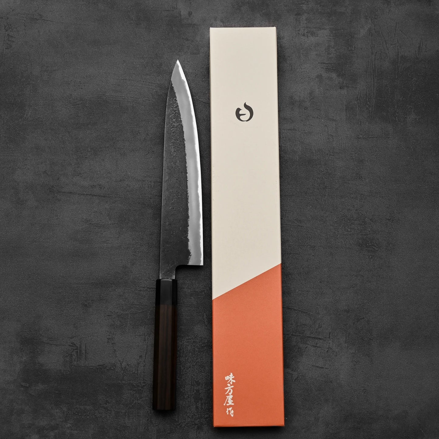 A Hinoura kurouchi nashiji AS (stainless clad) gyuto 240mm kitchen knife with a textured blade and dark handle by Hinoura sits next to a cream and orange rectangular box on a dark textured surface.