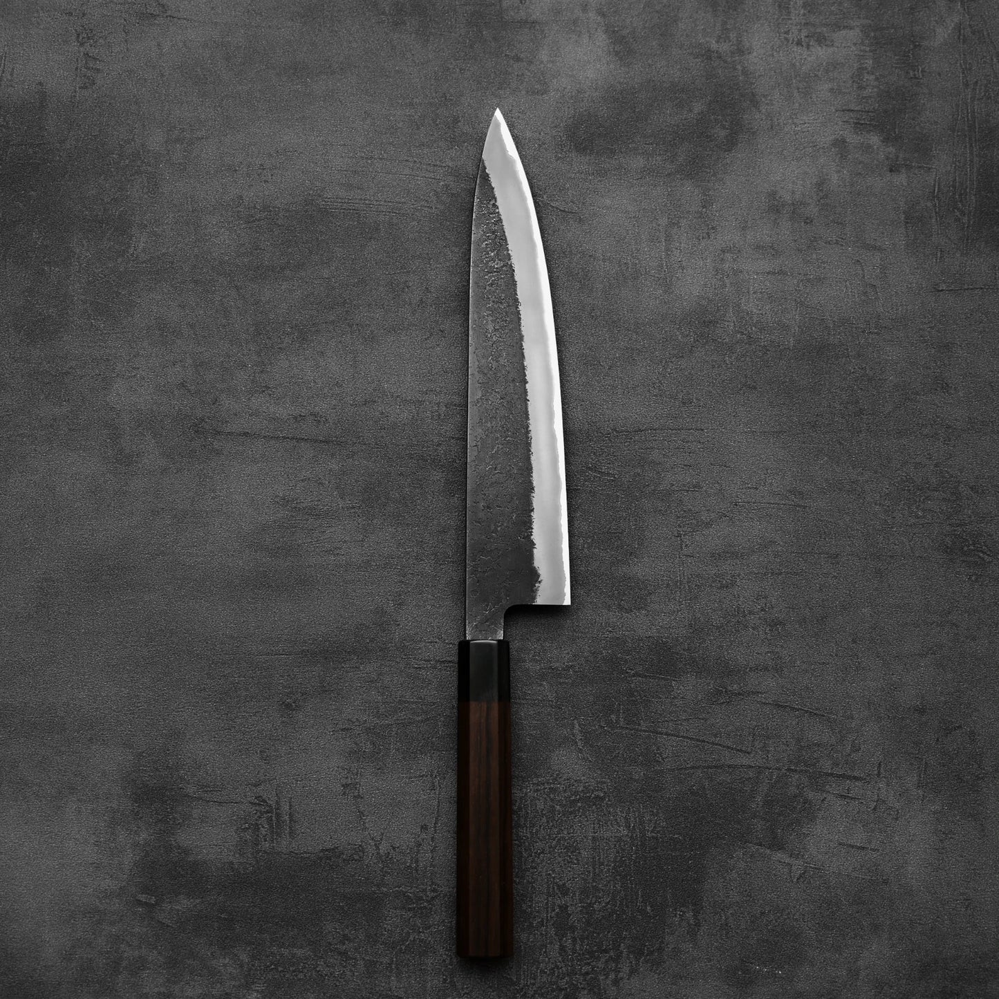 The Hinoura kurouchi nashiji AS (stainless clad) gyuto 240mm by Hinoura, featuring a dark wooden handle and textured blade, lies flat on a dark gray, textured surface.