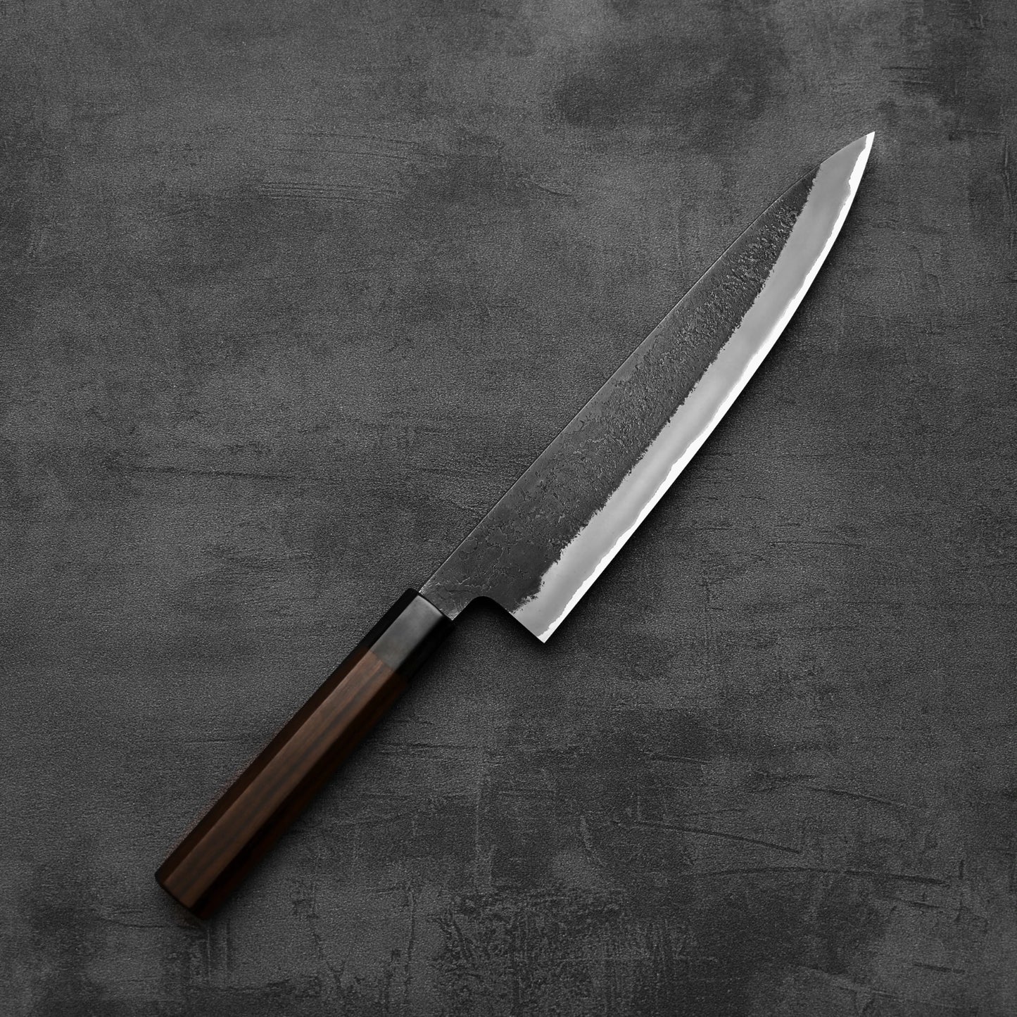 A Hinoura kurouchi nashiji AS (stainless clad) gyuto 240mm kitchen knife by Hinoura, featuring a dark wooden handle and a shiny, textured blade, rests on a gray, textured surface.