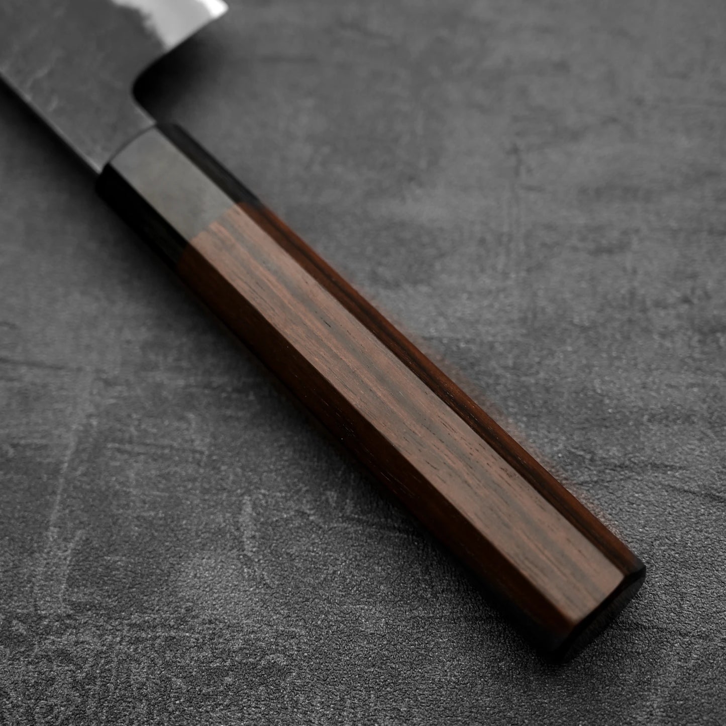 Close-up of the Hinoura kurouchi nashiji AS (stainless clad) gyuto 240mm by Hinoura, a Japanese kitchen knife with a dark wooden handle on a textured dark gray surface. The Aogami Super steel blade is partially visible in the upper left corner.