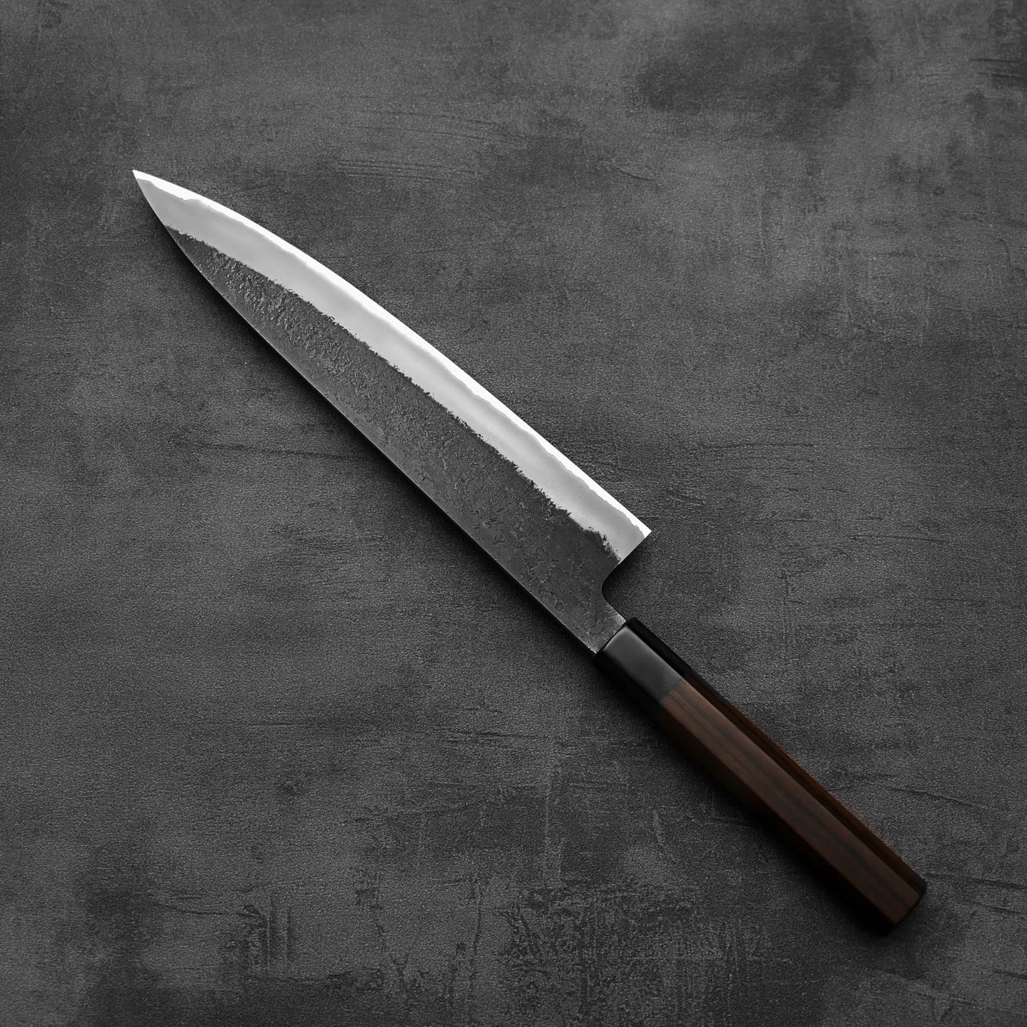 A Hinoura kurouchi nashiji AS (stainless clad) gyuto 240mm by Hinoura, featuring a dark wooden handle and a rustic, hammered finish near the spine, rests on a textured surface. The blade is crafted from Aogami Super steel with a polished edge.