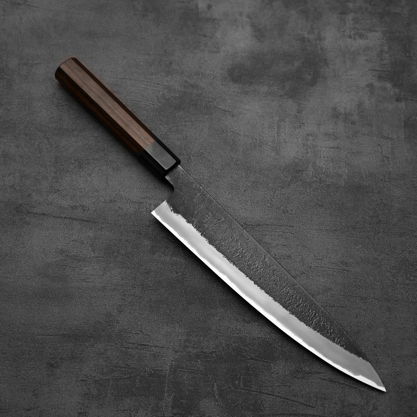 A Hinoura kurouchi nashiji AS (stainless clad) gyuto 240mm by Hinoura, featuring a dark wooden handle and patterned Aogami Super steel blade, rests on a textured dark gray surface.