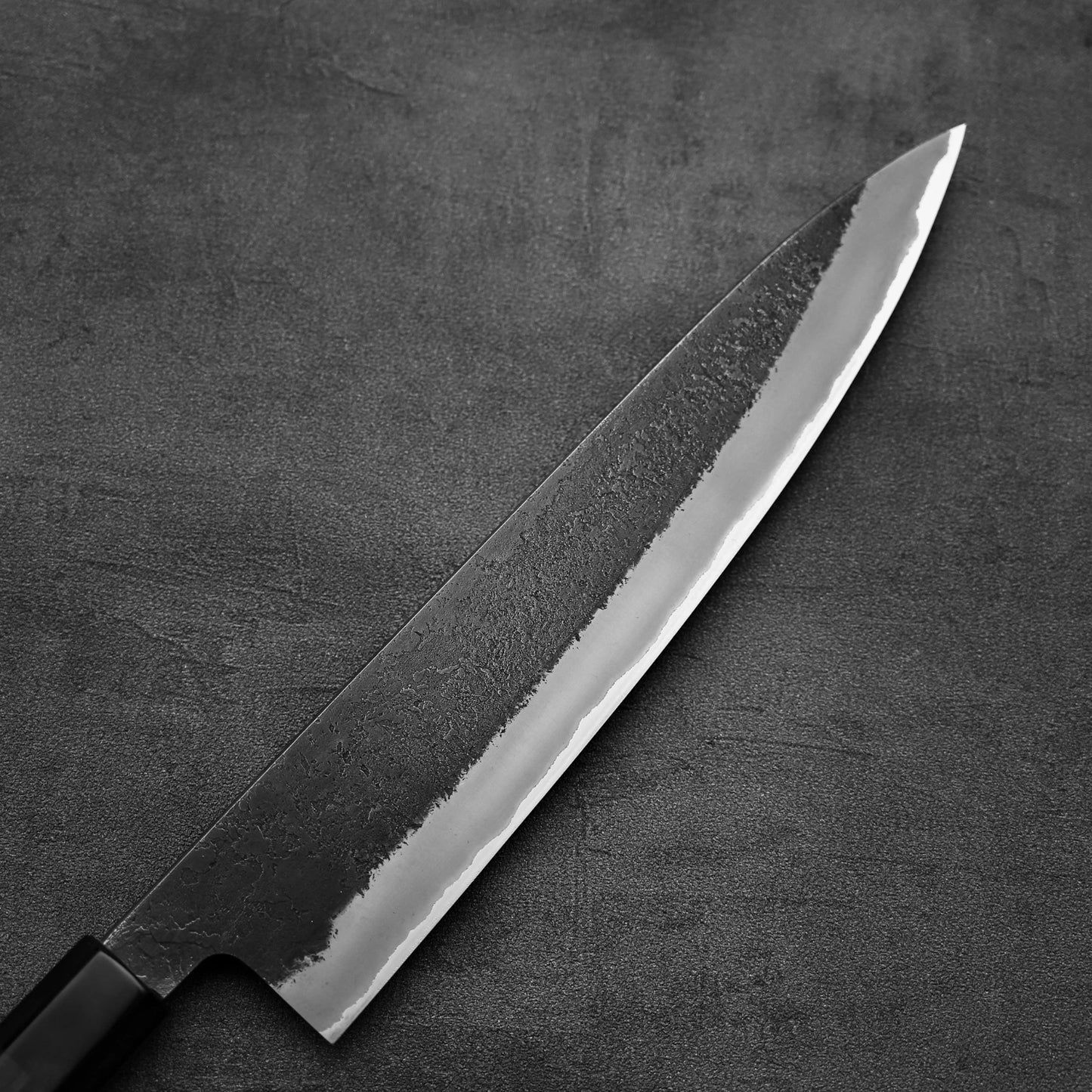 A close-up of the Hinoura kurouchi nashiji AS (stainless clad) gyuto 240mm by Hinoura, featuring a sharp, polished Aogami Super steel blade and dark handle on a textured gray surface.