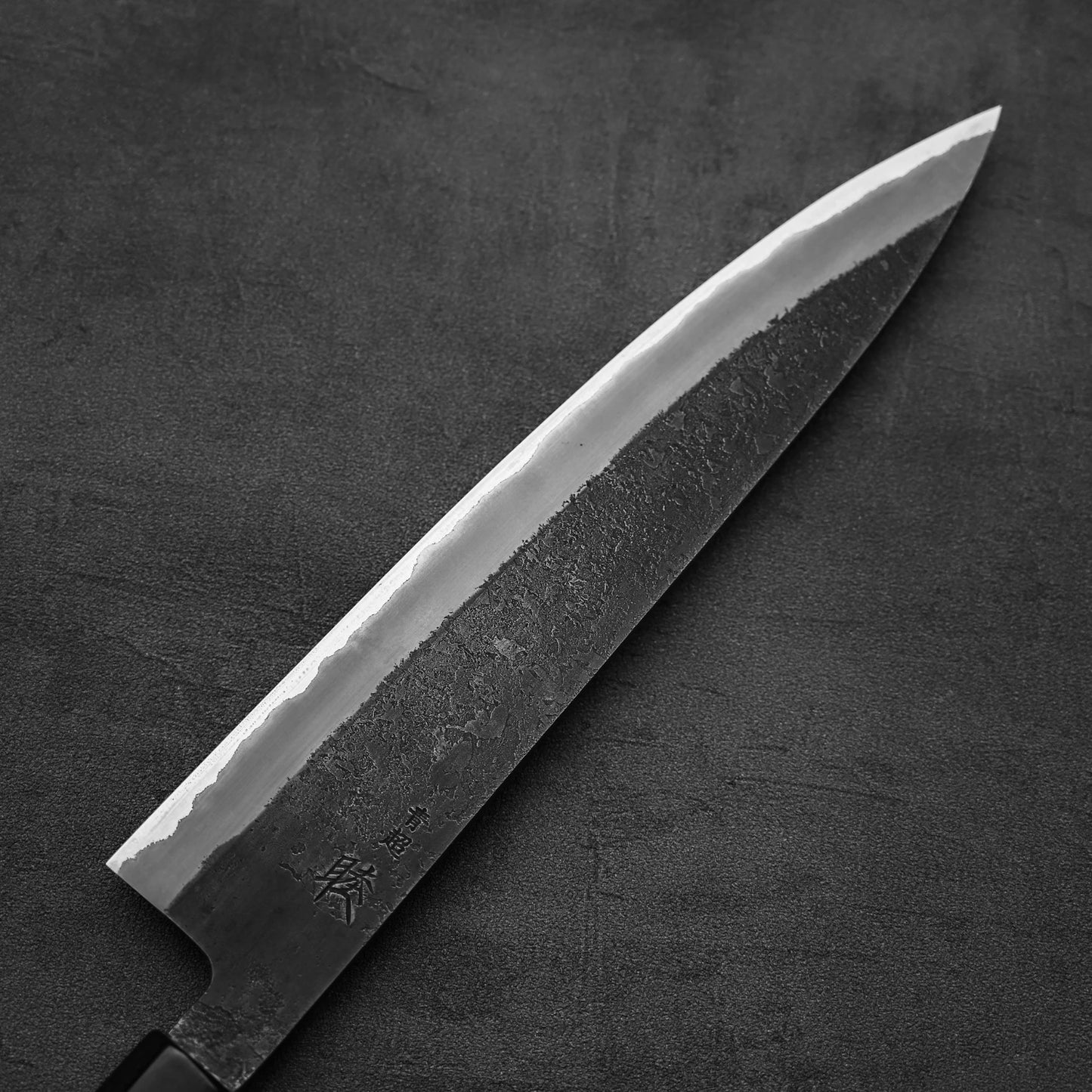 A close-up of the Hinoura kurouchi nashiji AS (stainless clad) gyuto 240mm by Hinoura, featuring a textured dark blade and sharp edge, lying on a dark surface.
