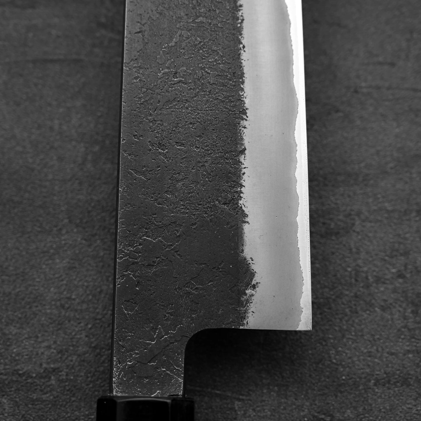Close-up of the Hinoura kurouchi nashiji AS (stainless clad) gyuto 240mm blade, highlighting its dark kurouchi finish near the spine, smooth shiny edge, and partially visible handle against a dark textured background.