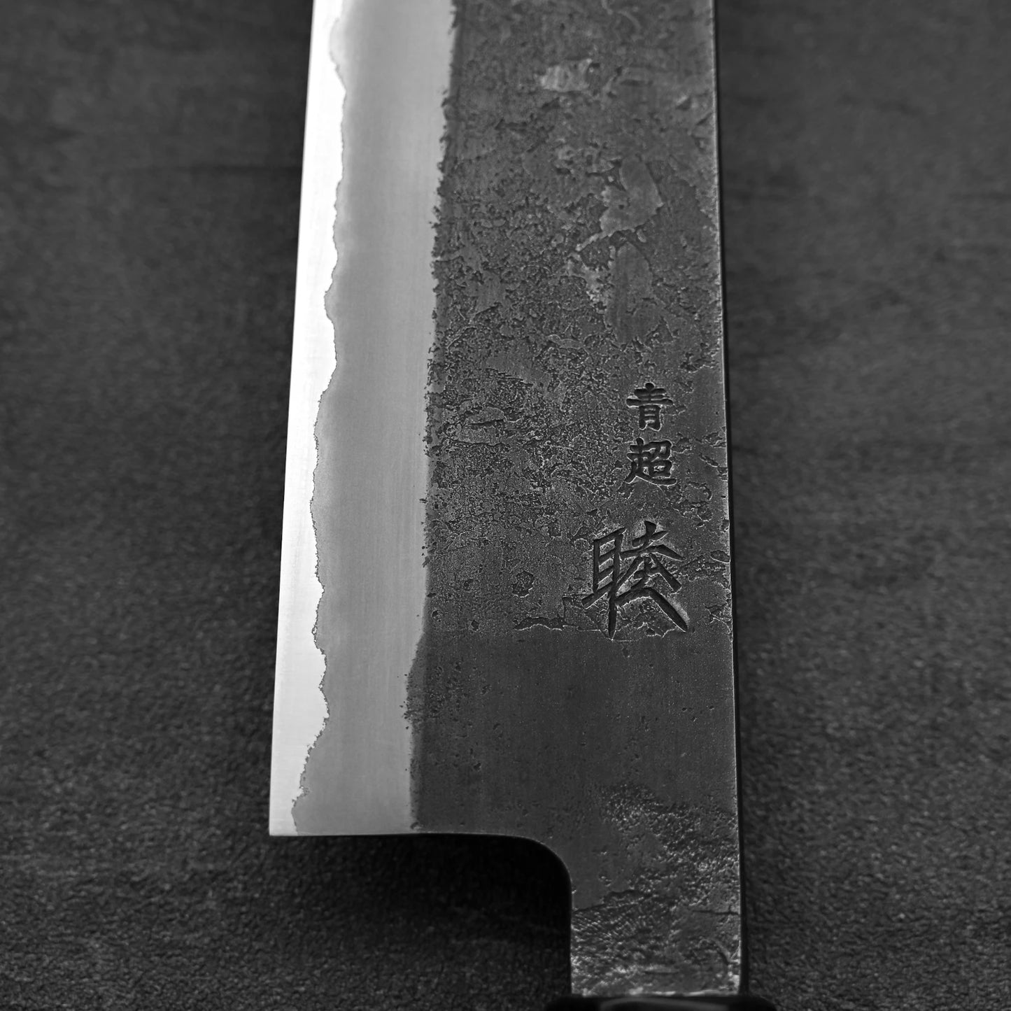 A close-up of the Hinoura kurouchi nashiji AS (stainless clad) gyuto 240mm blade shows textured steel and engraved Japanese characters, highlighting the craftsmanship of this Hinoura kitchen knife against a dark background.