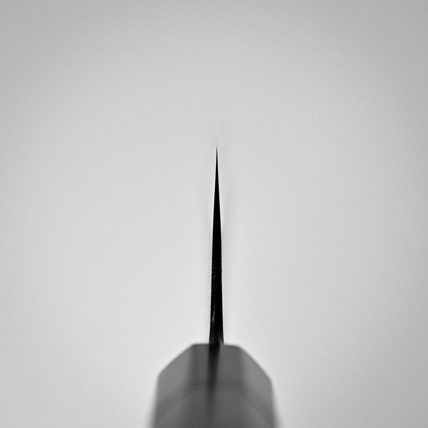 Close-up, centered view of the pointed tip of the Hinoura kurouchi nashiji AS (stainless clad) gyuto 240mm, highlighting the sharpness and precision of this Japanese kitchen knifes blade against a plain light gray background.