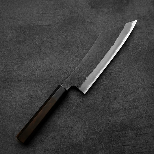 The Hinoura kurouchi nashiji AS (stainless clad) hakata 180mm knife—with its dark handle and wide, angled blade—rests on a textured dark gray surface, highlighting the craftsmanship of Hinoura.