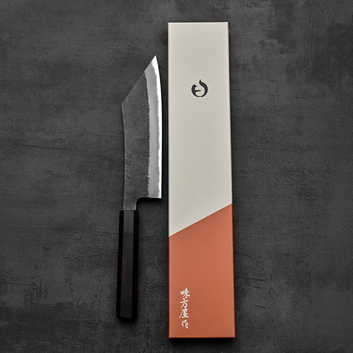 The Hinoura kurouchi nashiji AS (stainless clad) hakata 180mm, a handmade Japanese knife with a dark handle, rests beside its beige and rust-orange box with Japanese characters and a logo, all on a dark textured surface.
