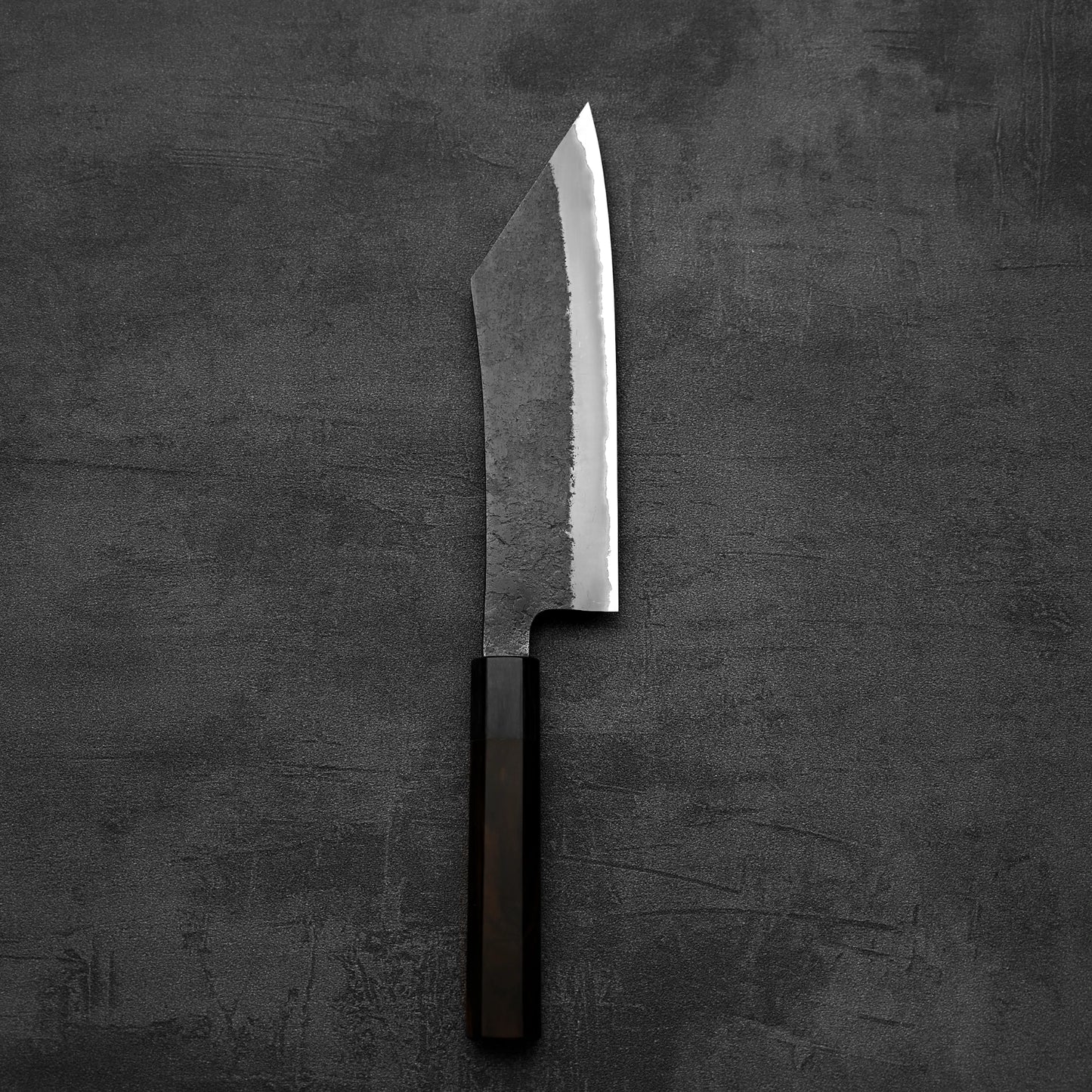 The Hinoura kurouchi nashiji AS hakata 180mm—a large handmade Japanese knife with a dark wooden handle and wide Aogami Super steel blade—rests flat on a dark, textured surface.