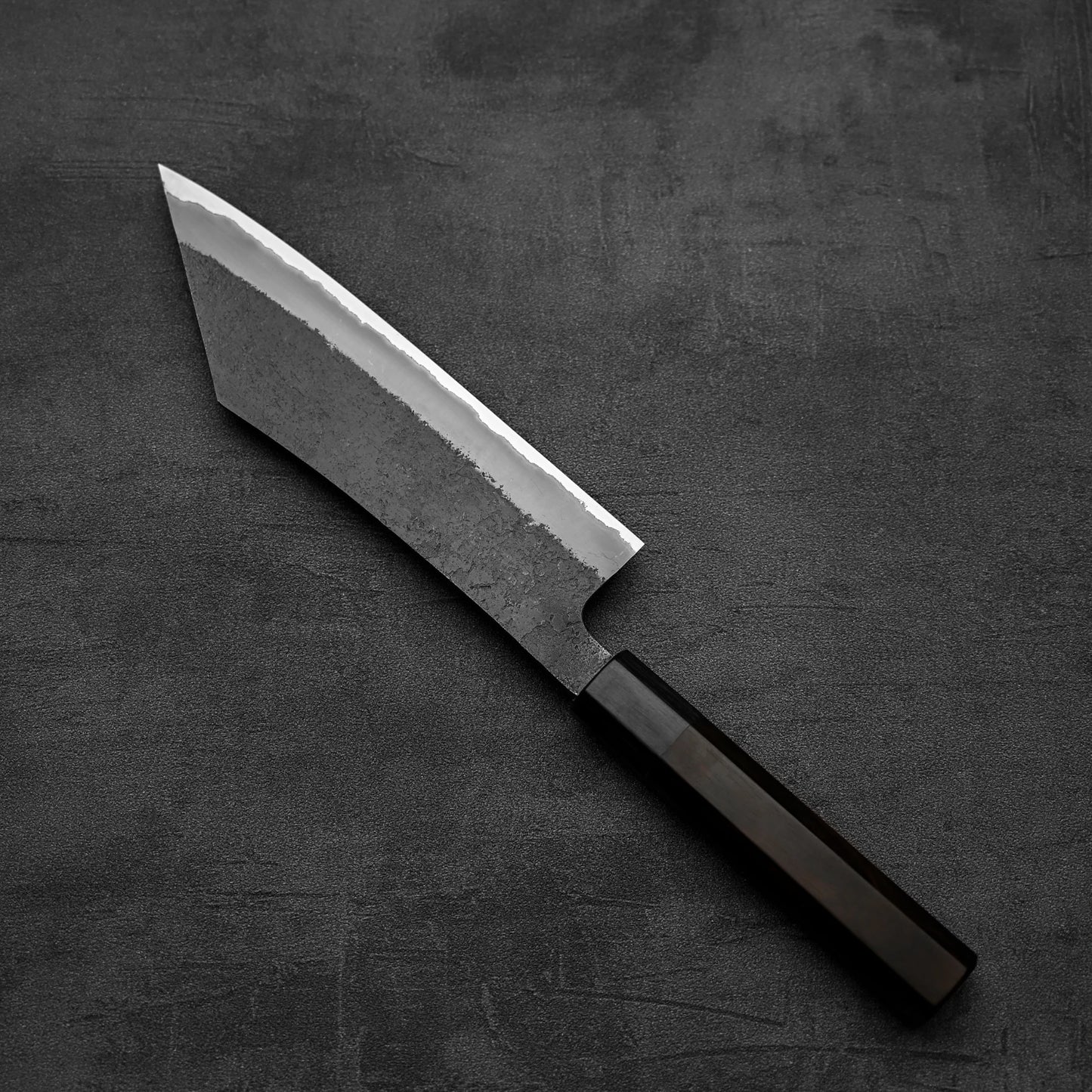 The Hinoura kurouchi nashiji AS (stainless clad) hakata 180mm knife by Hinoura, featuring a dark handle and wide, angular blade of Aogami Super steel, rests on a textured dark gray surface.