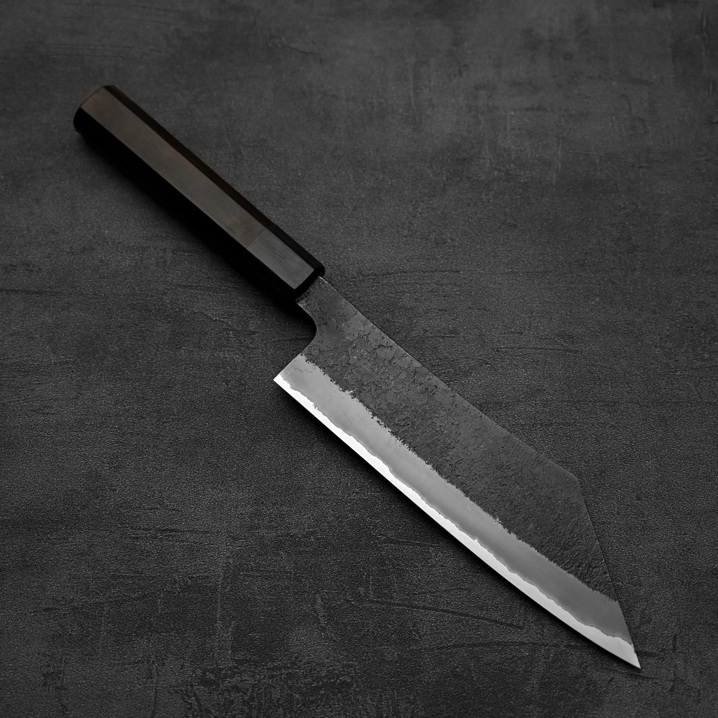 The Hinoura kurouchi nashiji AS (stainless clad) hakata 180mm knife, featuring a dark wooden handle and a textured Aogami Super steel blade, rests on a dark, textured surface.