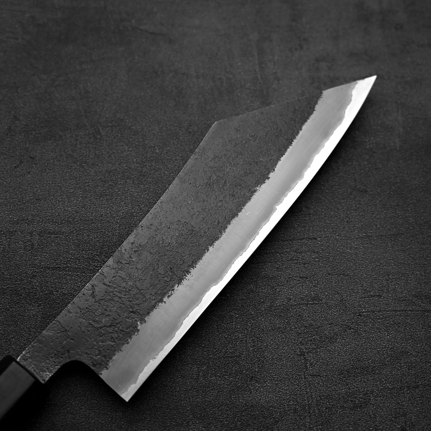 A close-up of the Hinoura kurouchi nashiji AS (stainless clad) hakata 180mm knife by Hinoura, showcasing its sharp, angular blade and textured dark finish against a dark backdrop.