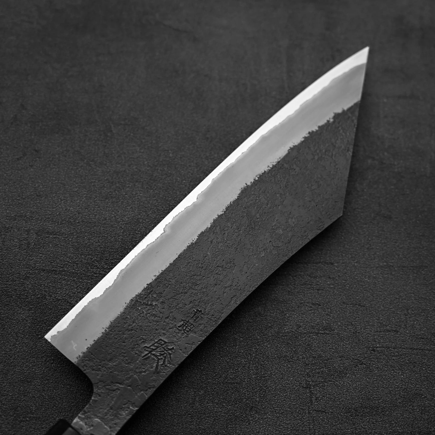 A close-up of the Hinoura kurouchi nashiji AS (stainless clad) hakata 180mm Japanese knife, featuring a textured blade with engraved markings, crafted from Aogami Super steel, by Hinoura, shown on a dark, textured surface.