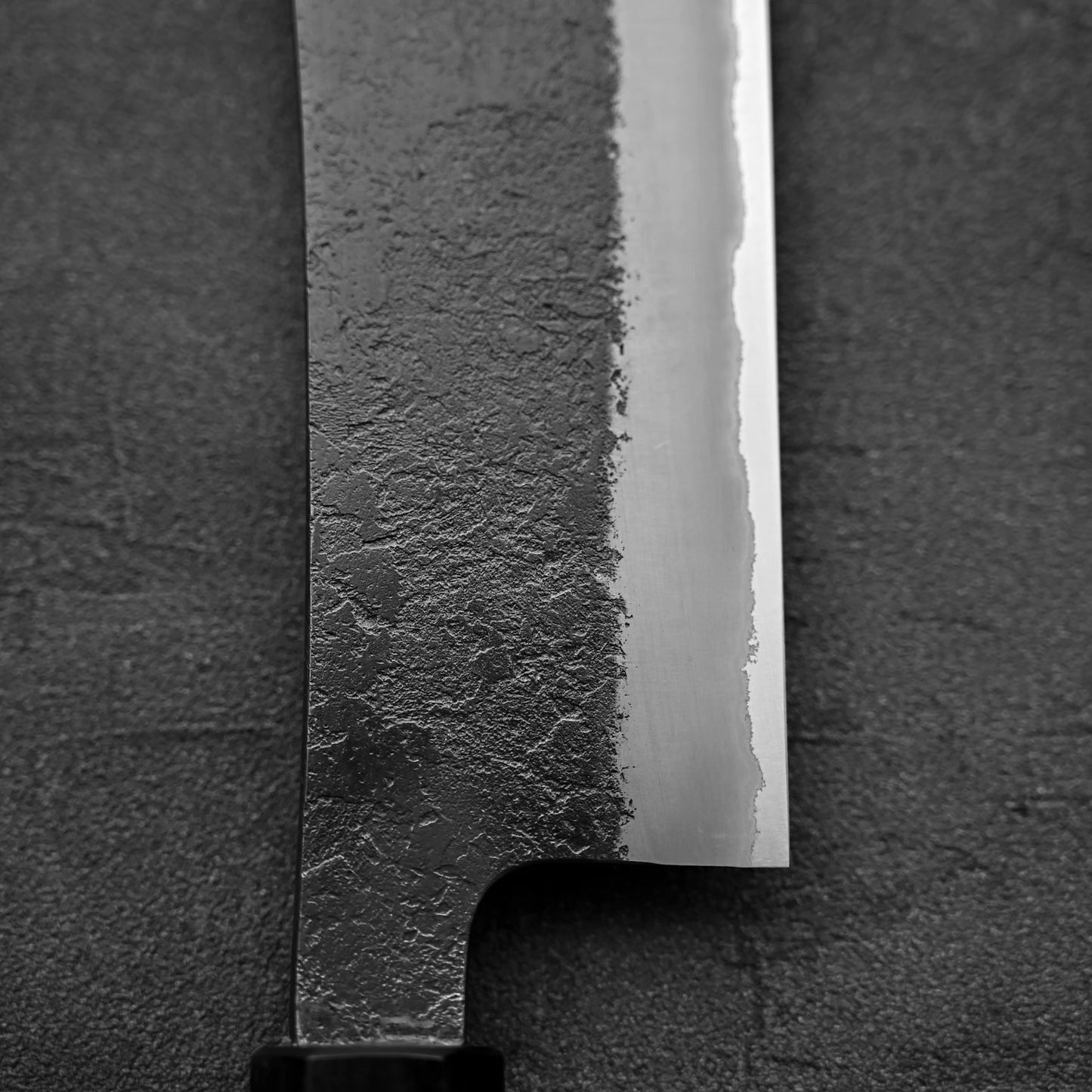 A close-up of the Hinoura kurouchi nashiji AS (stainless clad) hakata 180mm knife displays its textured dark finish and polished edge, highlighting Hinoura craftsmanship against a dark background.