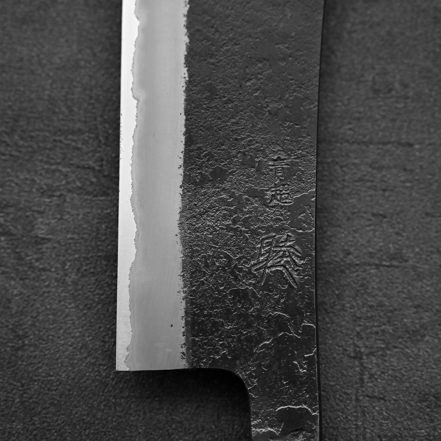 Close-up of the Hinoura kurouchi nashiji AS (stainless clad) hakata 180mm blade, showcasing a striking contrast between polished and rough steel textures, Japanese engraving near the edge, set against a dark background. Brand: Hinoura.