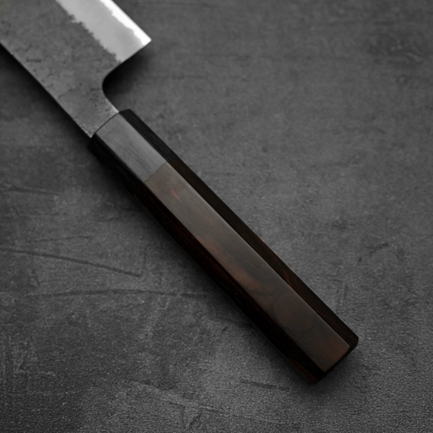 A close-up of the Hinoura kurouchi nashiji AS (stainless clad) hakata 180mm by Hinoura, showing the dark polished wood handle and part of the blade, set against a dark textured background.