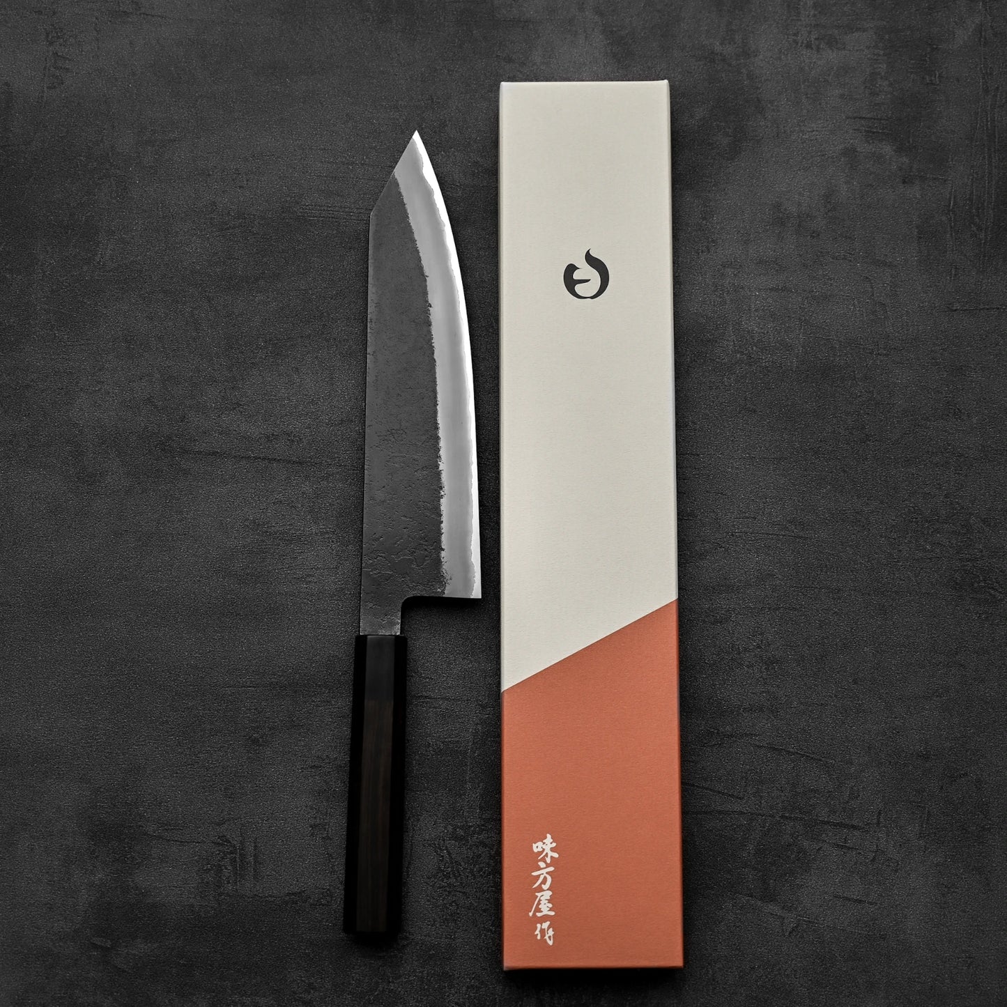 The Hinoura kurouchi nashiji AS (stainless clad) kiritsuke gyuto 210mm by Hinoura, with a dark handle, rests beside a minimalist box with Japanese characters on a dark textured background—crafted in the famed Hinoura style.