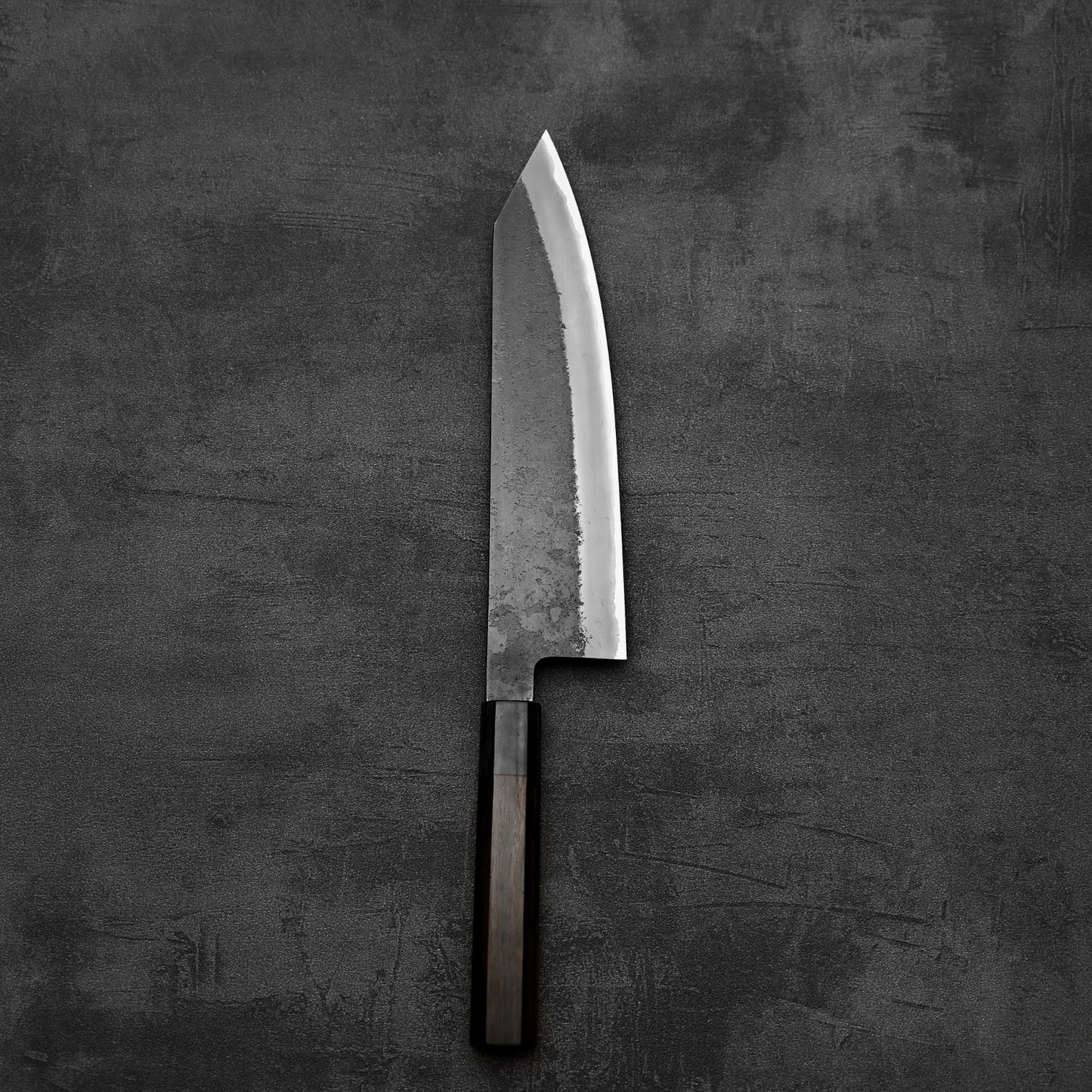 A Hinoura kurouchi nashiji AS (stainless clad) kiritsuke gyuto 210mm chef’s knife with a dark wooden handle and sharp blade lies flat on a textured dark gray surface.