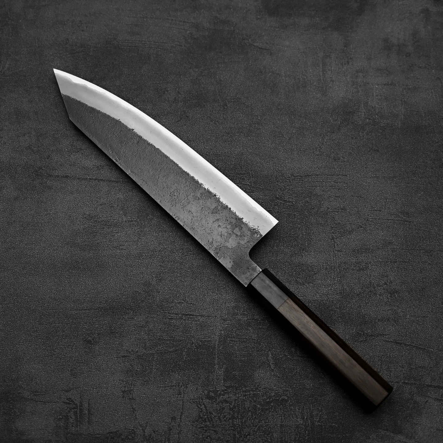 A Hinoura kurouchi nashiji AS (stainless clad) kiritsuke gyuto 210mm kitchen knife with a textured blade and dark handle from Hinoura rests on a dark, textured surface.