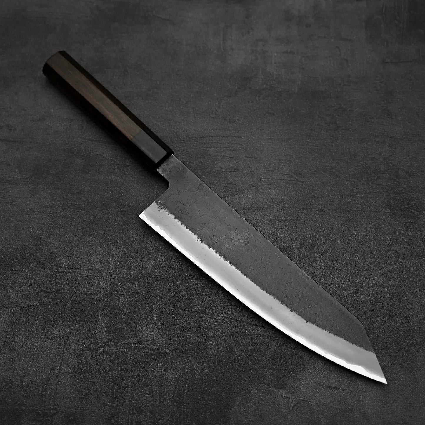 A Hinoura kurouchi nashiji AS (stainless clad) kiritsuke gyuto 210mm kitchen knife by Hinoura with a dark wooden handle and sharp rectangular blade rests on a textured dark gray surface.