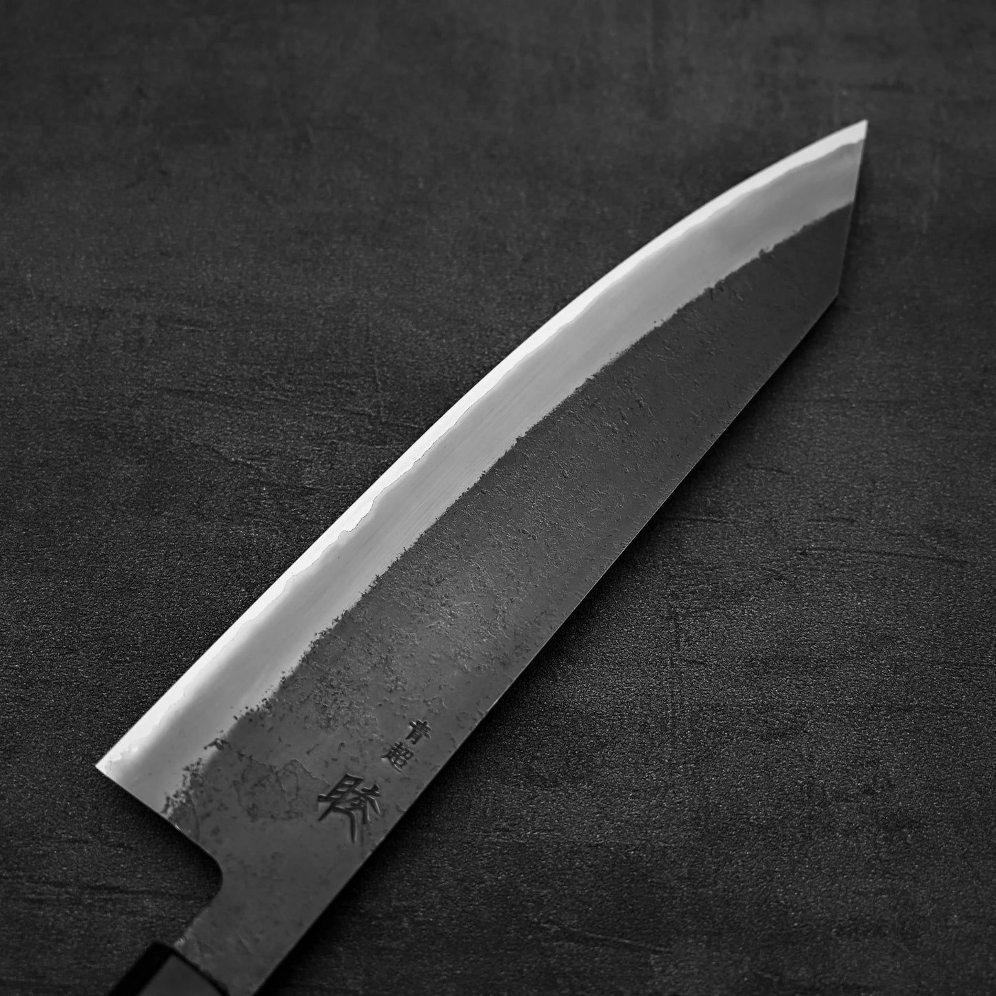 A close-up of the Hinoura kurouchi nashiji AS (stainless clad) kiritsuke gyuto 210mm by Hinoura, showing its sharp, angled Aogami Super steel blade with kanji characters, set on a dark matte surface.
