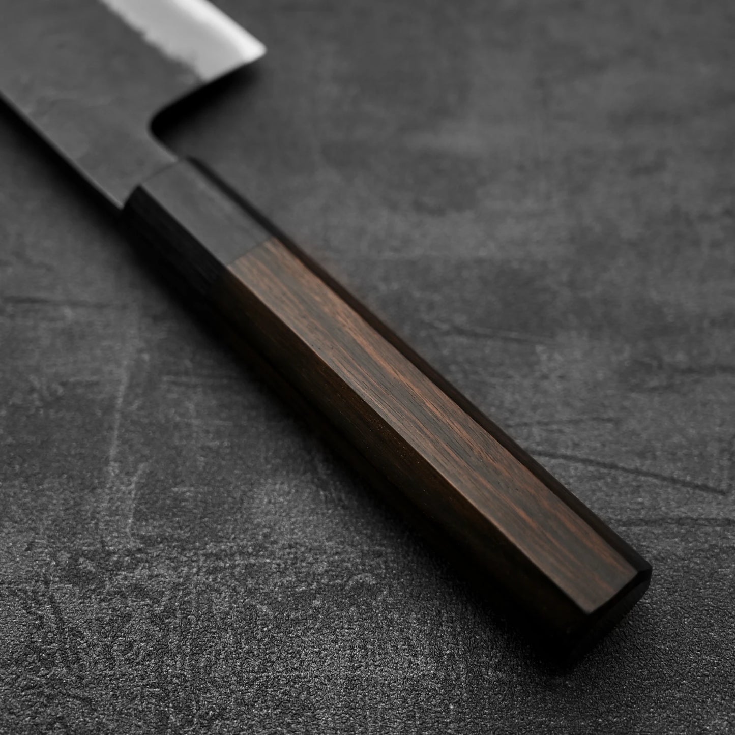 A close-up of a Hinoura kurouchi nashiji AS (stainless clad) kiritsuke gyuto 210mm, showing the wooden handle and partial blade, rests on a textured dark gray surface. The knife is crafted by Hinoura.