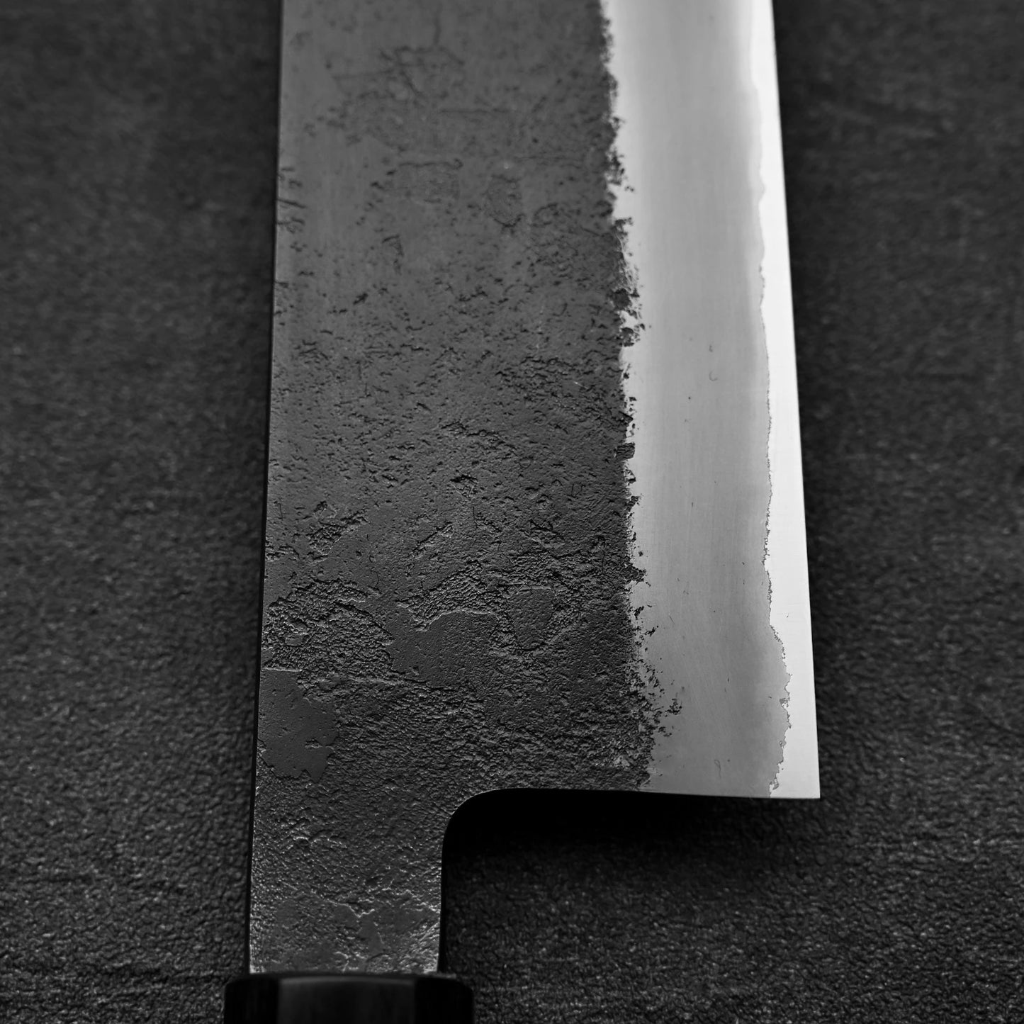 Close-up of a Hinoura kurouchi nashiji AS (stainless clad) kiritsuke gyuto 210mm blade by Hinoura, highlighting its textured dark forged finish that transitions to a polished edge, set against a dark background.