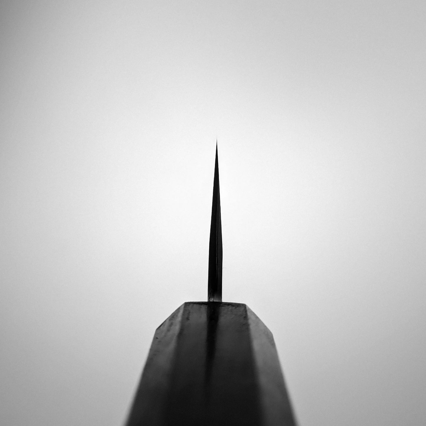 Close-up of the Nakagawa aogami#1 damascus bunka 180mm - etched by Satoshi Nakagawa, showing its razor-sharp, thin carbon steel blade tip pointing upward against a light gray background, highlighting its precision and craftsmanship.
