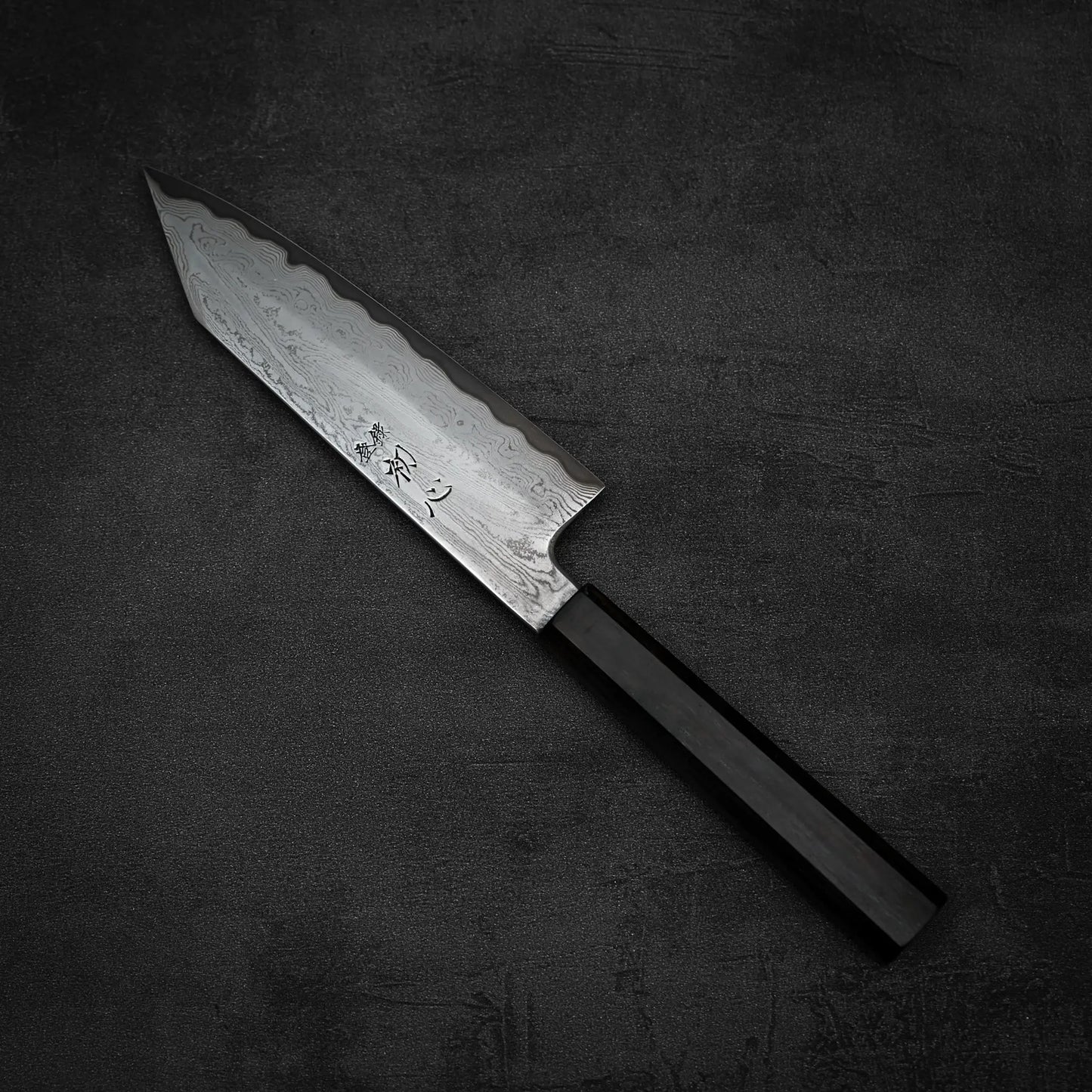 A Satoshi Nakagawa Nakagawa aogami#1 damascus bunka 180mm – etched knife with a patterned Damascus blade and dark, angular handle rests on a textured surface, highlighting the elegance of Japanese carbon steel craftsmanship.