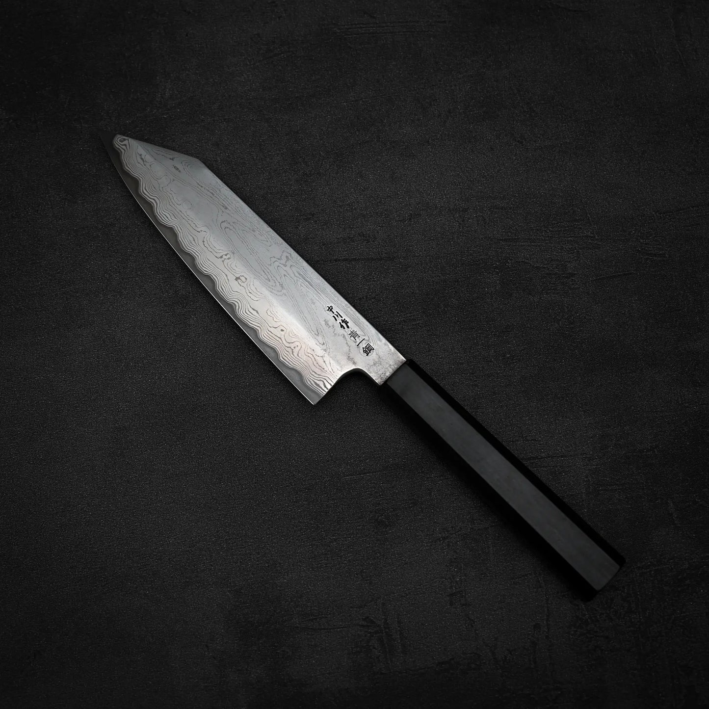 A Satoshi Nakagawa aogami#1 damascus bunka 180mm - etched knife with a patterned blade and black handle rests on a dark, textured surface.