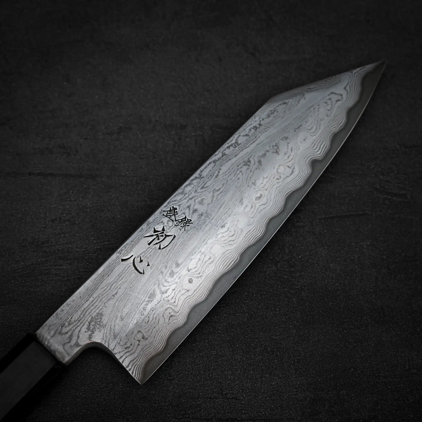 A close-up of the Satoshi Nakagawa Nakagawa aogami#1 damascus bunka 180mm - etched knife, showcasing its patterned Damascus steel blade with Japanese characters, resting on a dark textured surface.