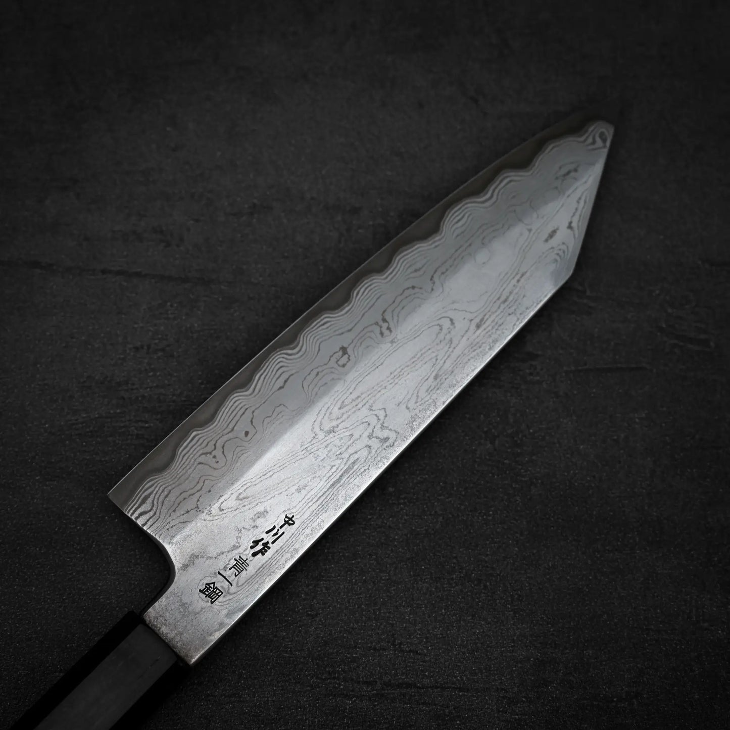 A close-up of the Satoshi Nakagawa Nakagawa aogami#1 damascus bunka 180mm - etched, featuring a patterned wavy blade with Japanese characters near the base and a black handle, resting on a dark textured surface.