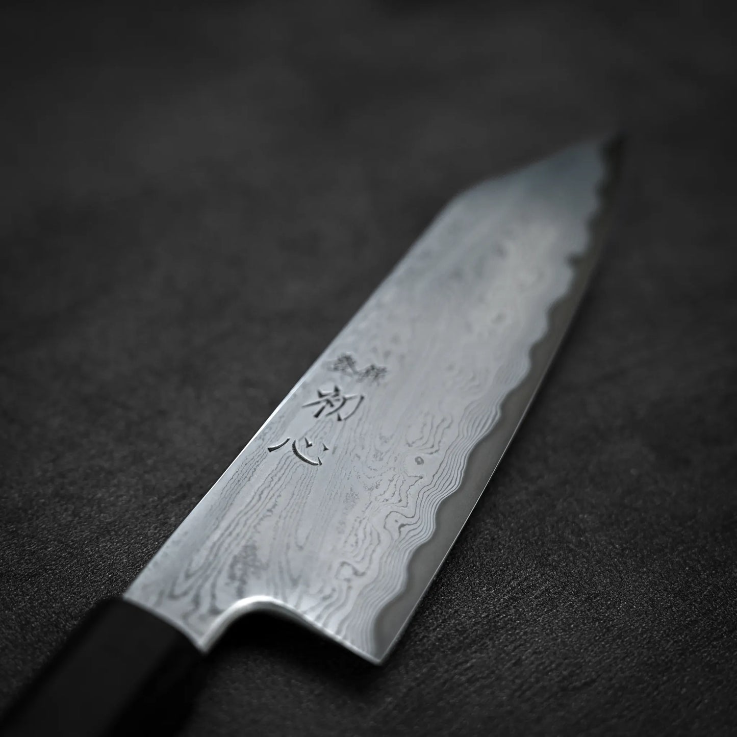 A close-up of the Satoshi Nakagawa Nakagawa aogami#1 damascus bunka 180mm - etched knife shows its wavy Damascus steel blade and engraved kanji, elegantly resting on a dark textured surface.