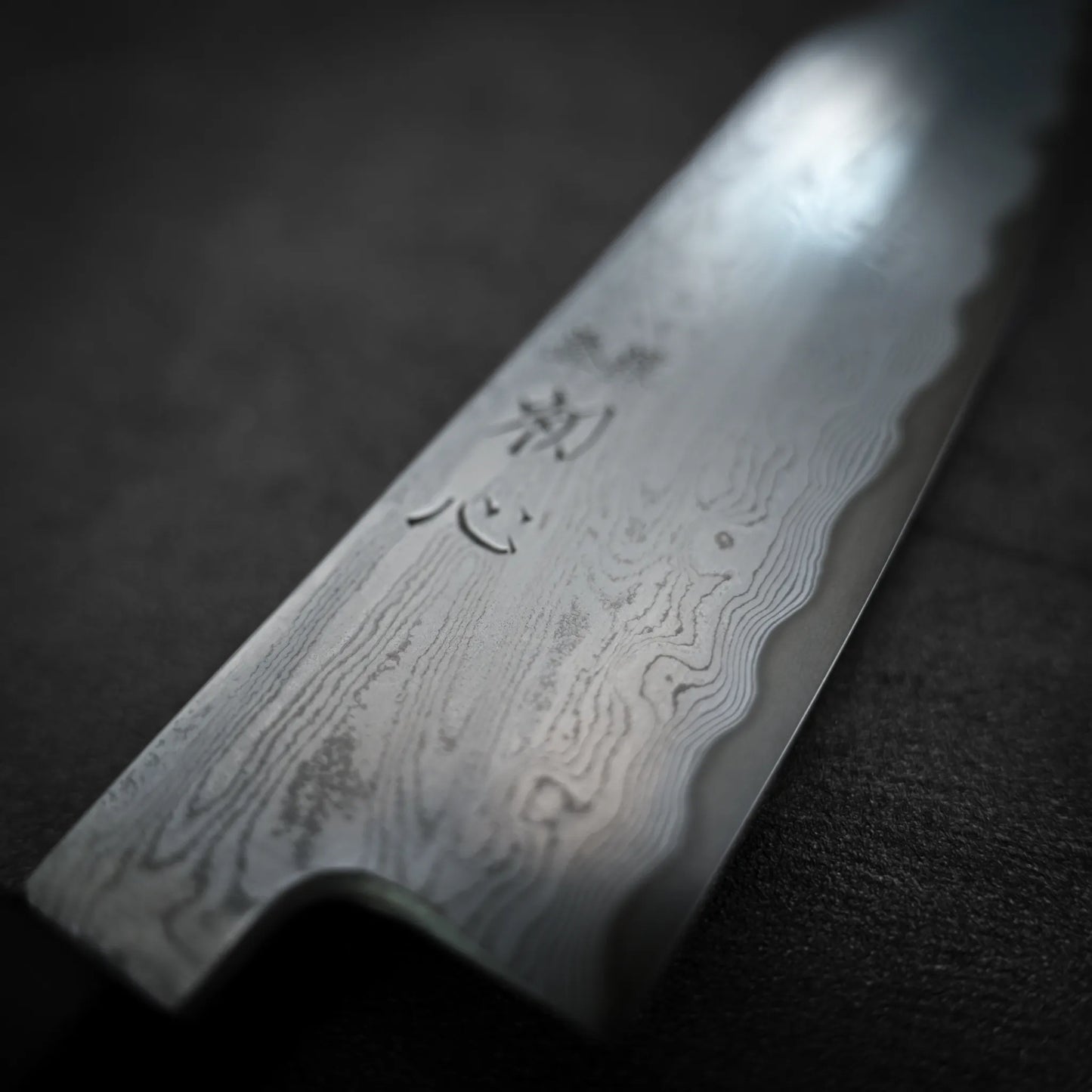 Close-up of the Satoshi Nakagawa Nakagawa aogami#1 damascus bunka 180mm - etched, featuring an intricate Damascus steel pattern and kanji engraving, highlighting the artistry of this Japanese carbon steel knife on a dark surface.