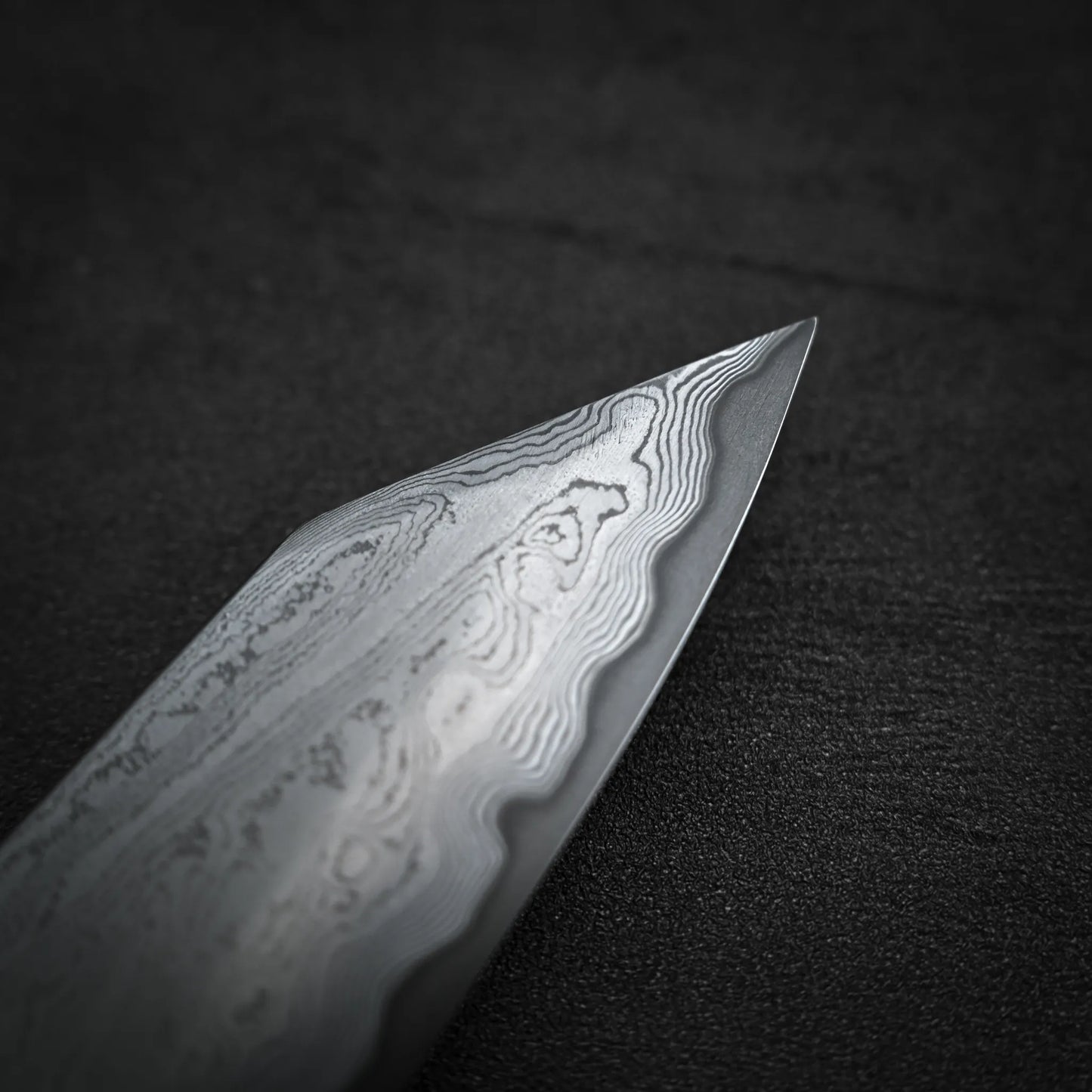 Close-up of the pointed tip of the Satoshi Nakagawa aogami#1 damascus bunka 180mm - etched, highlighting its unique wavy pattern against a dark, textured background.