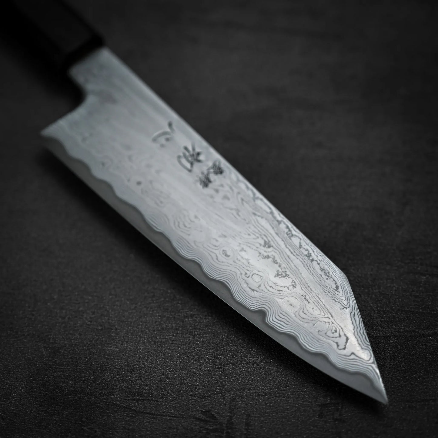 A close-up of the Satoshi Nakagawa Nakagawa aogami#1 damascus bunka 180mm - etched knife, displaying its dark handle and intricate Damascus wave patterns on the blade, set against a dark, textured background.