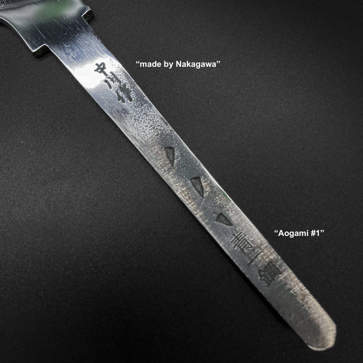 Close-up of the Satoshi Nakagawa aogami#1 honyaki tall gyuto 240mm blade, showing etched characters and English labels—“made by Nakagawa” and “Aogami #1 honyaki”—against a dark matte background.